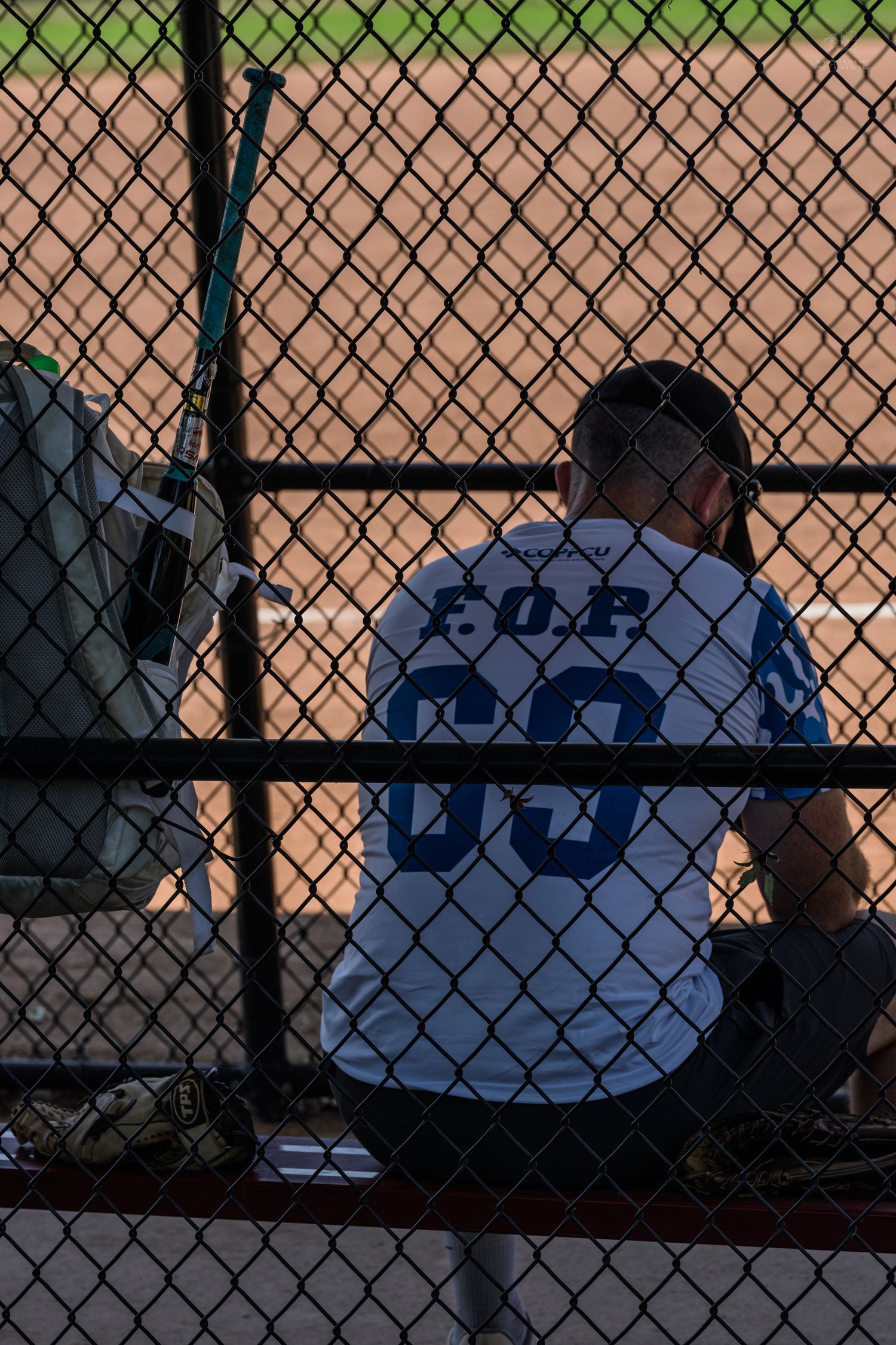 Player wearing FOP jersey number 00 sits in dugout behind chain-link fence at baseball field.