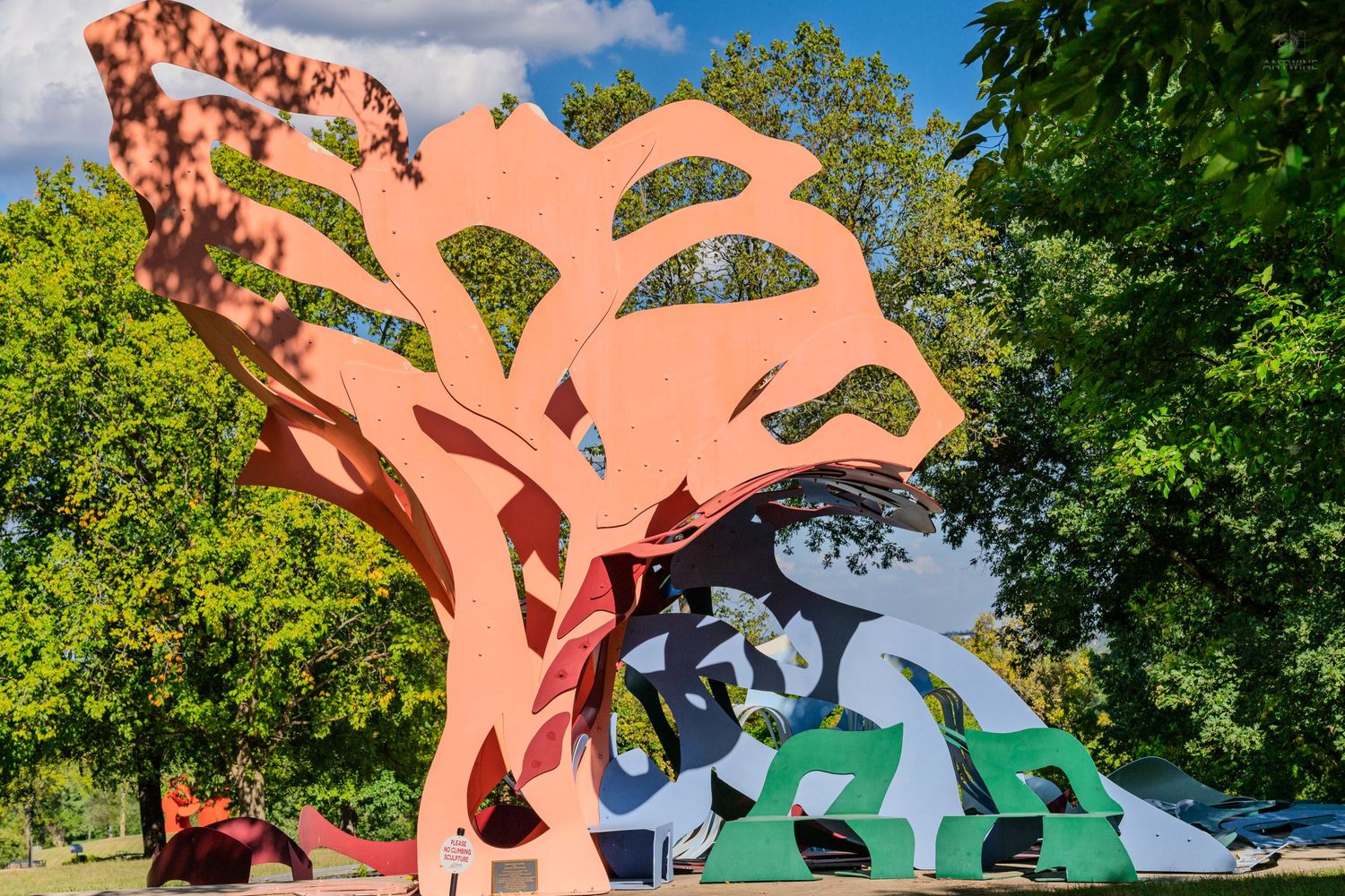 Colorful large-scale outdoor metal tree sculpture with abstract figures in a green park setting.