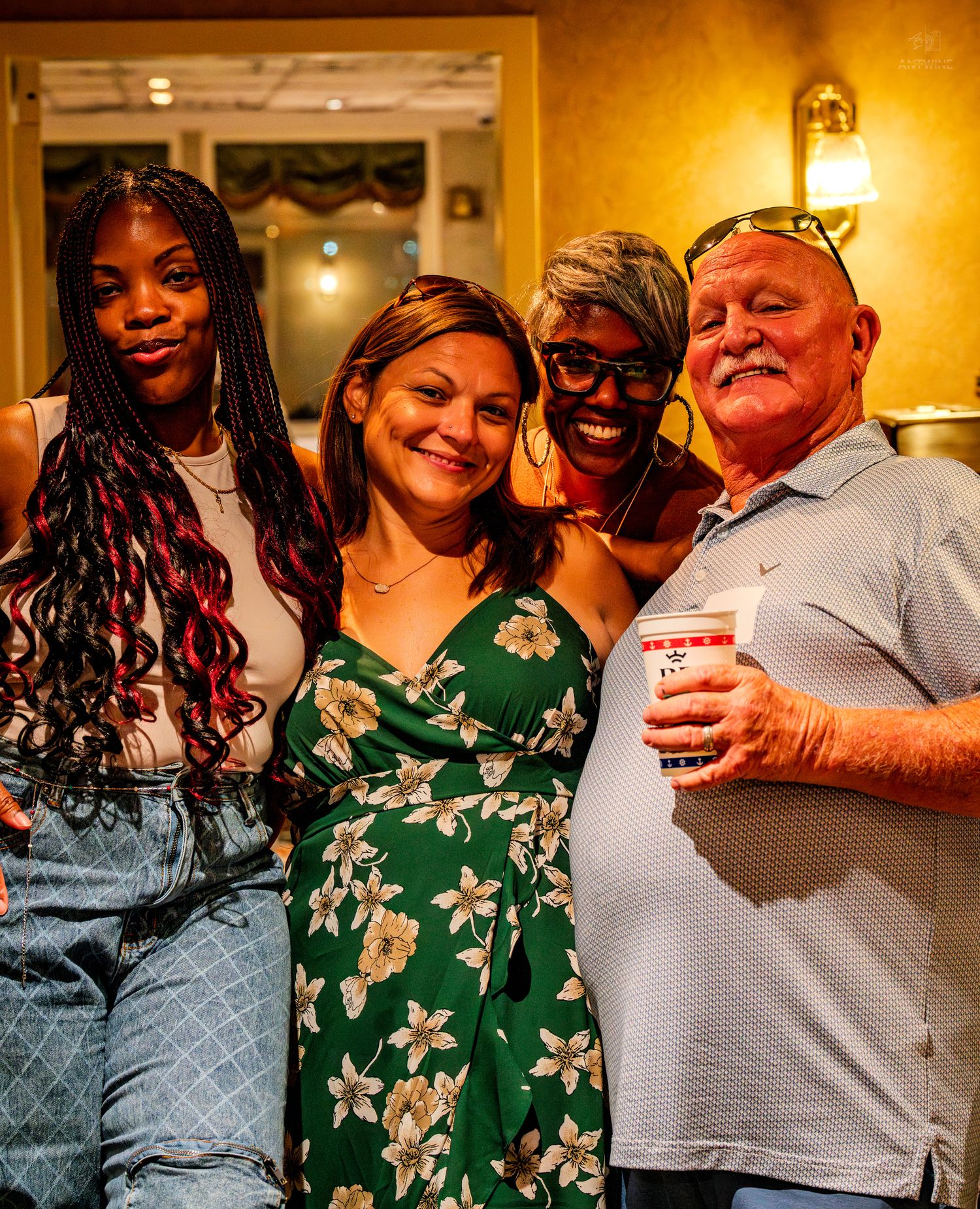 Four friends smiling together at an indoor gathering, including three women and one older man holding a drink.