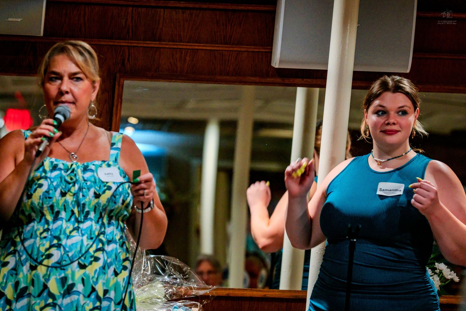 Two women on stage at an event, one speaking into a microphone in a floral dress, the other in blue.