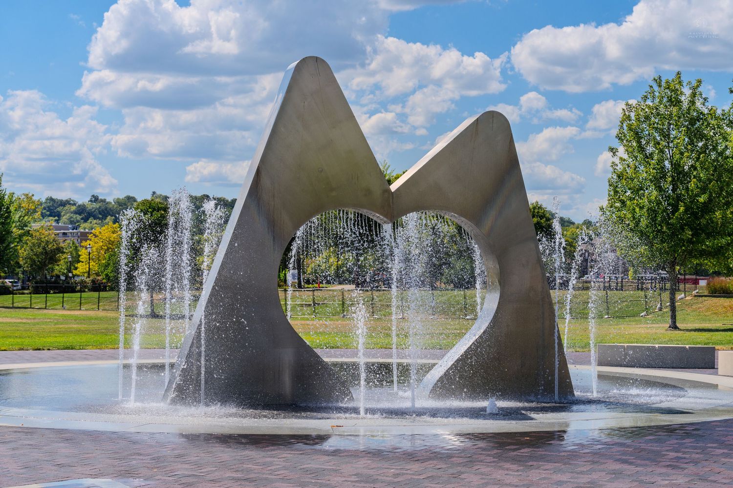 Modern abstract metal sculpture fountain with water jets in a sunny park setting.
