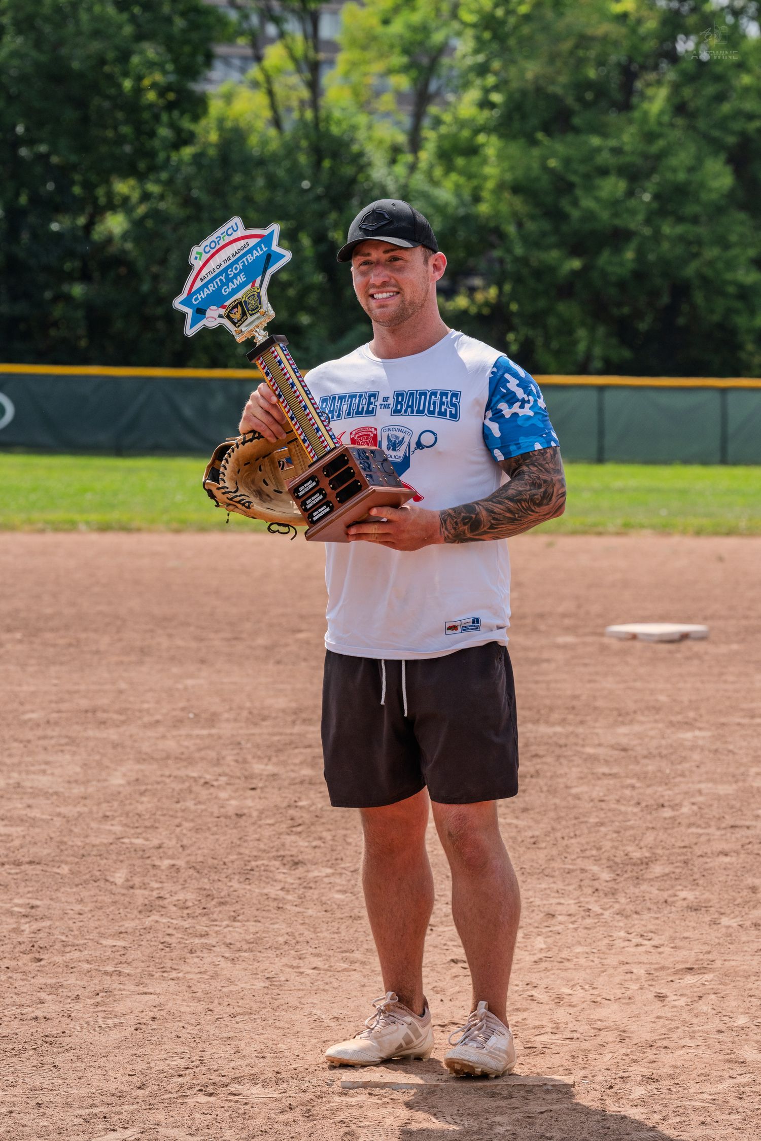 Man holding trophy on baseball field wearing Battle Badgers jersey and baseball glove on sunny day.