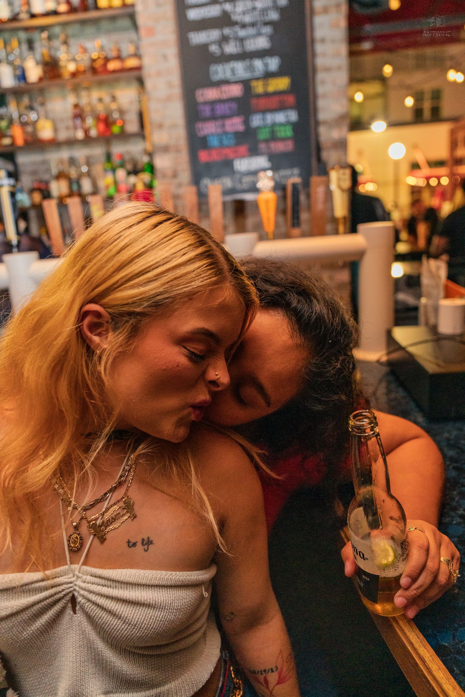Two women leaning close together at a bar, one holding a beer bottle.