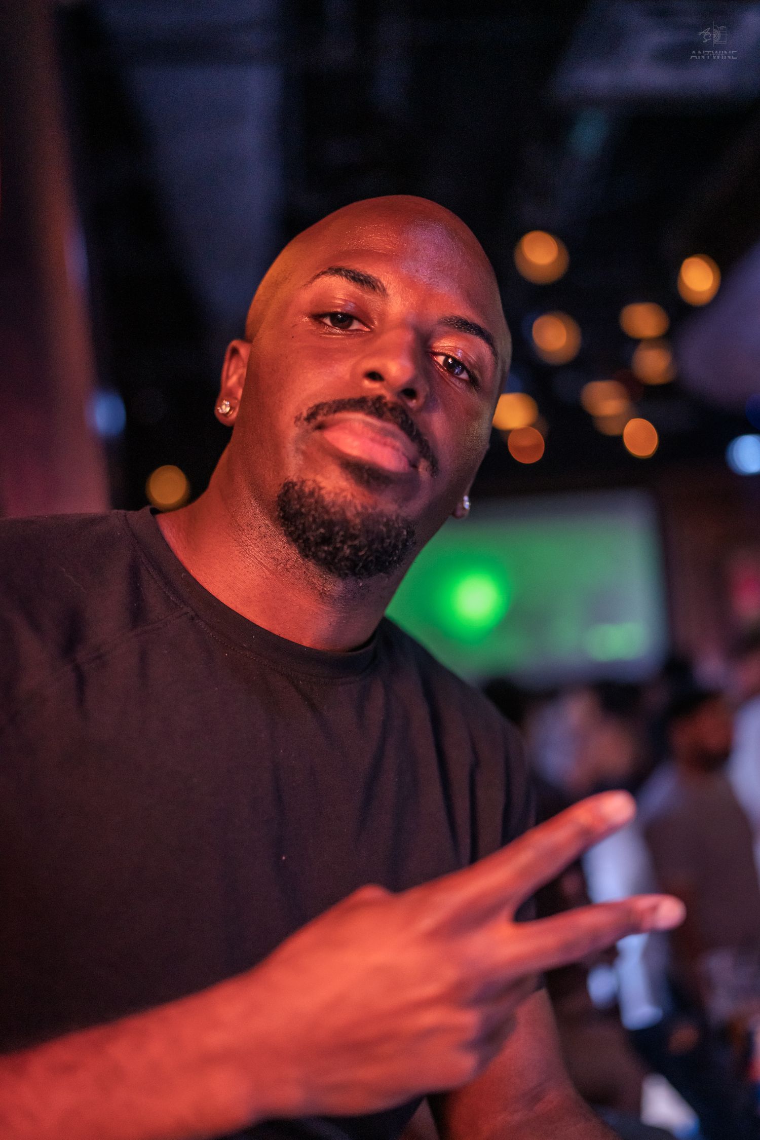 Bald Black man in black t-shirt smiles and flashes a peace sign under colorful nightclub lighting.