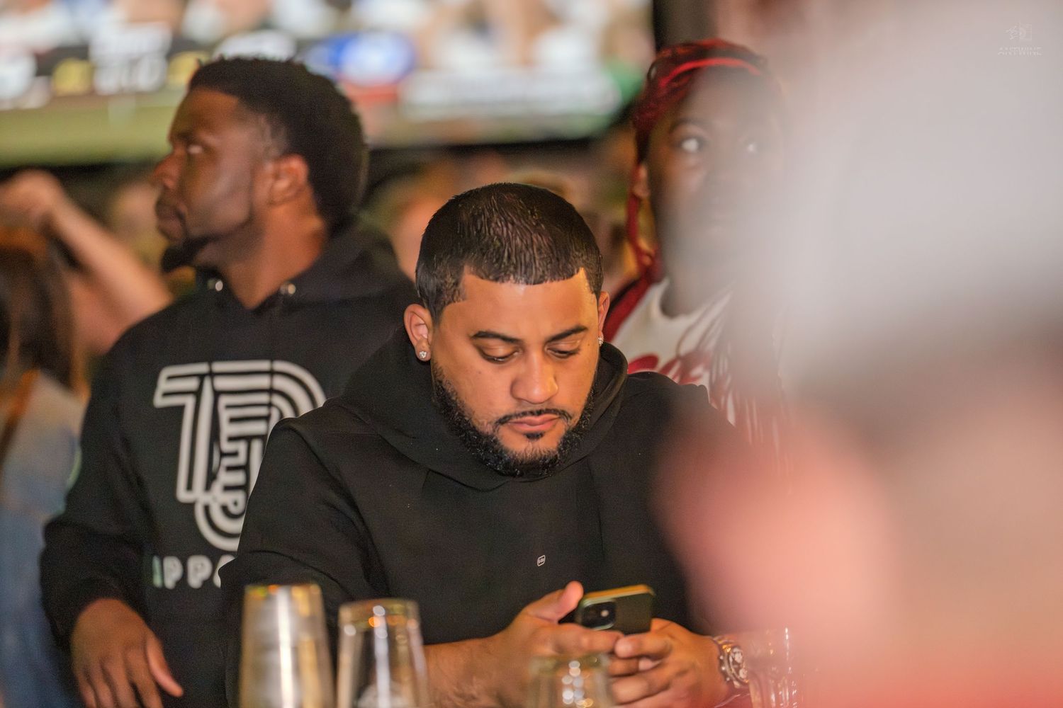 Man in black hoodie looking at his phone while sitting at a crowded bar or restaurant.
