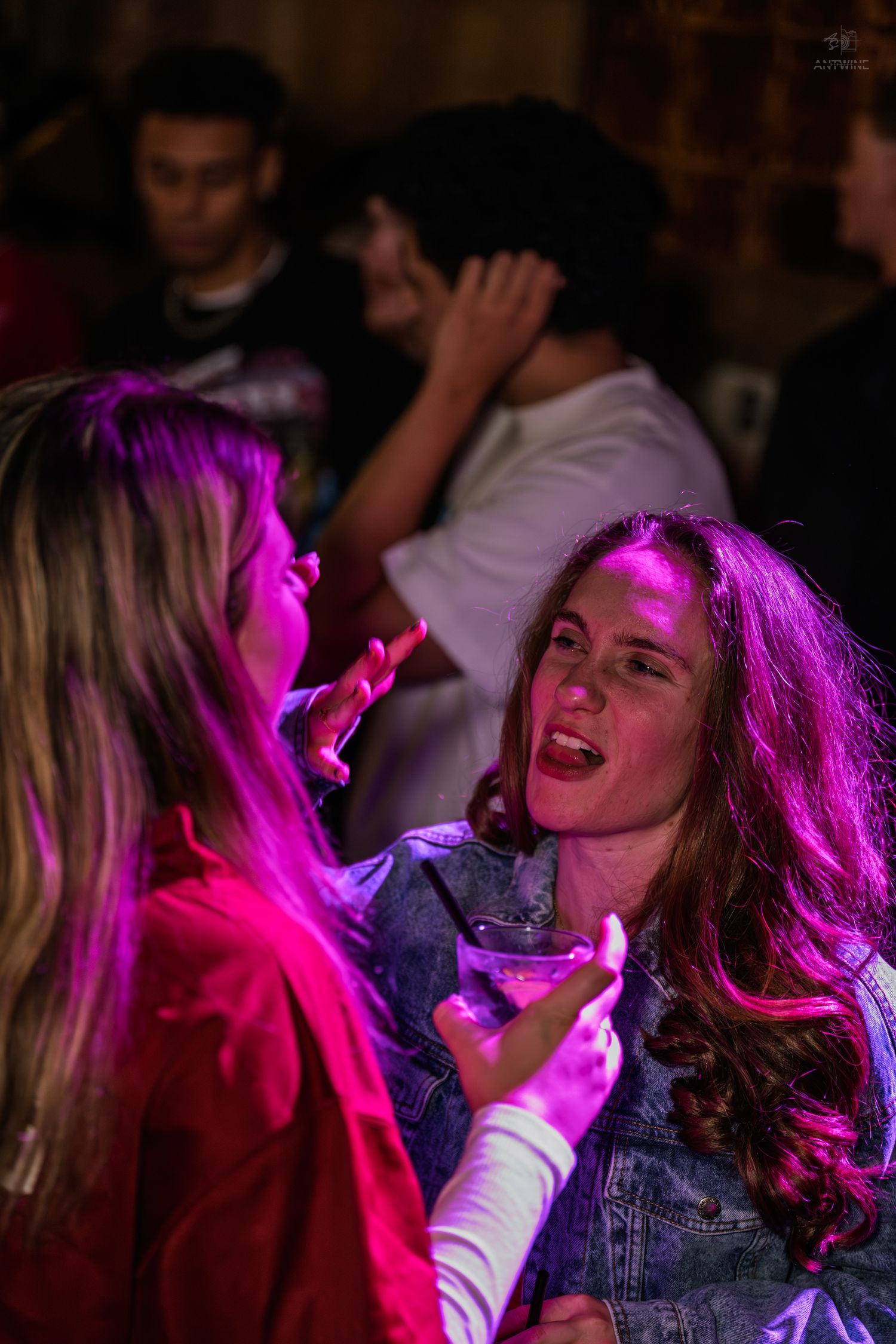 Two women laughing and talking under pink neon lights at a lively nightclub party.