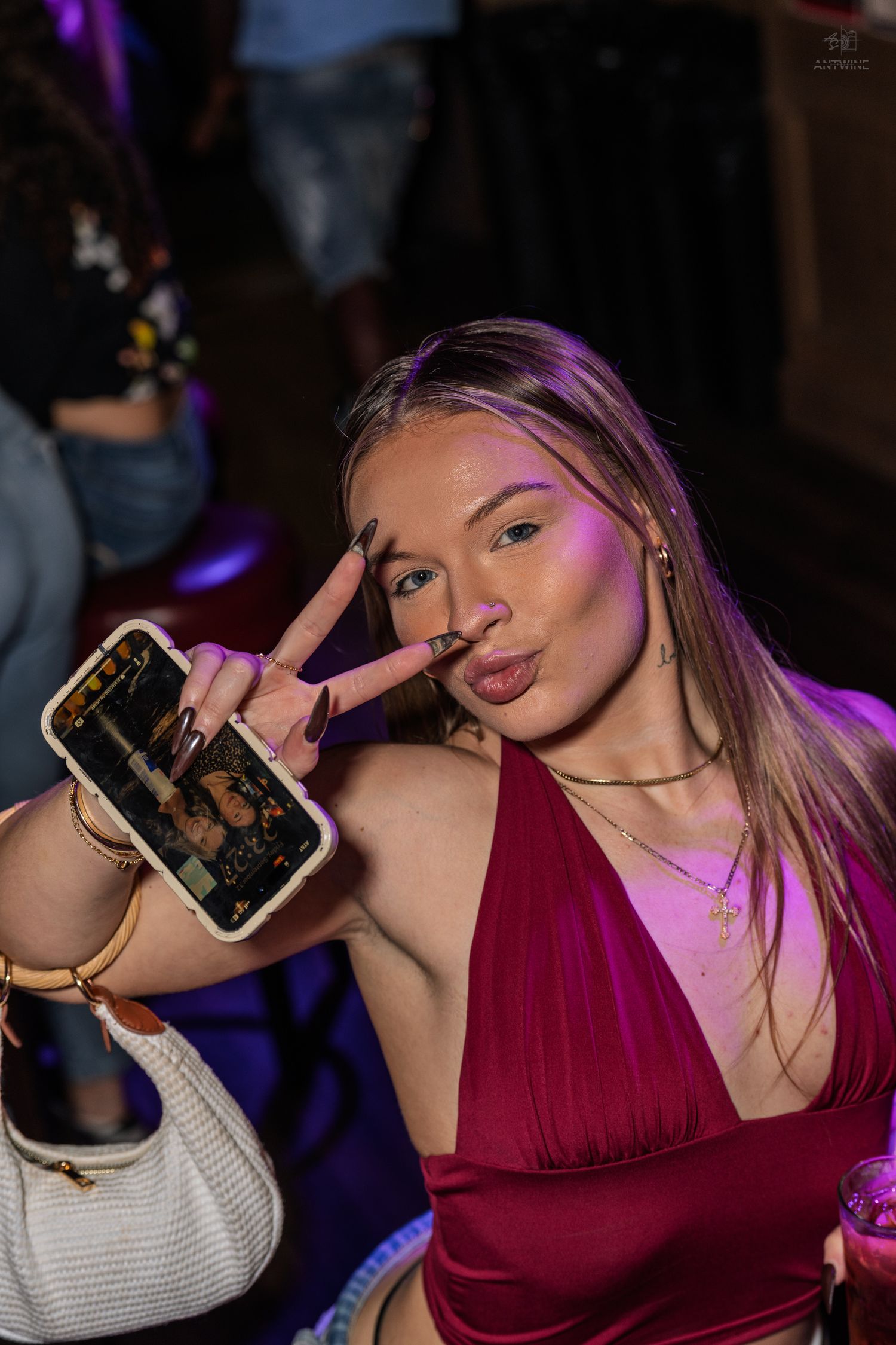 Blonde woman in burgundy dress poses with peace sign and pouty lips while holding phone at nightclub.