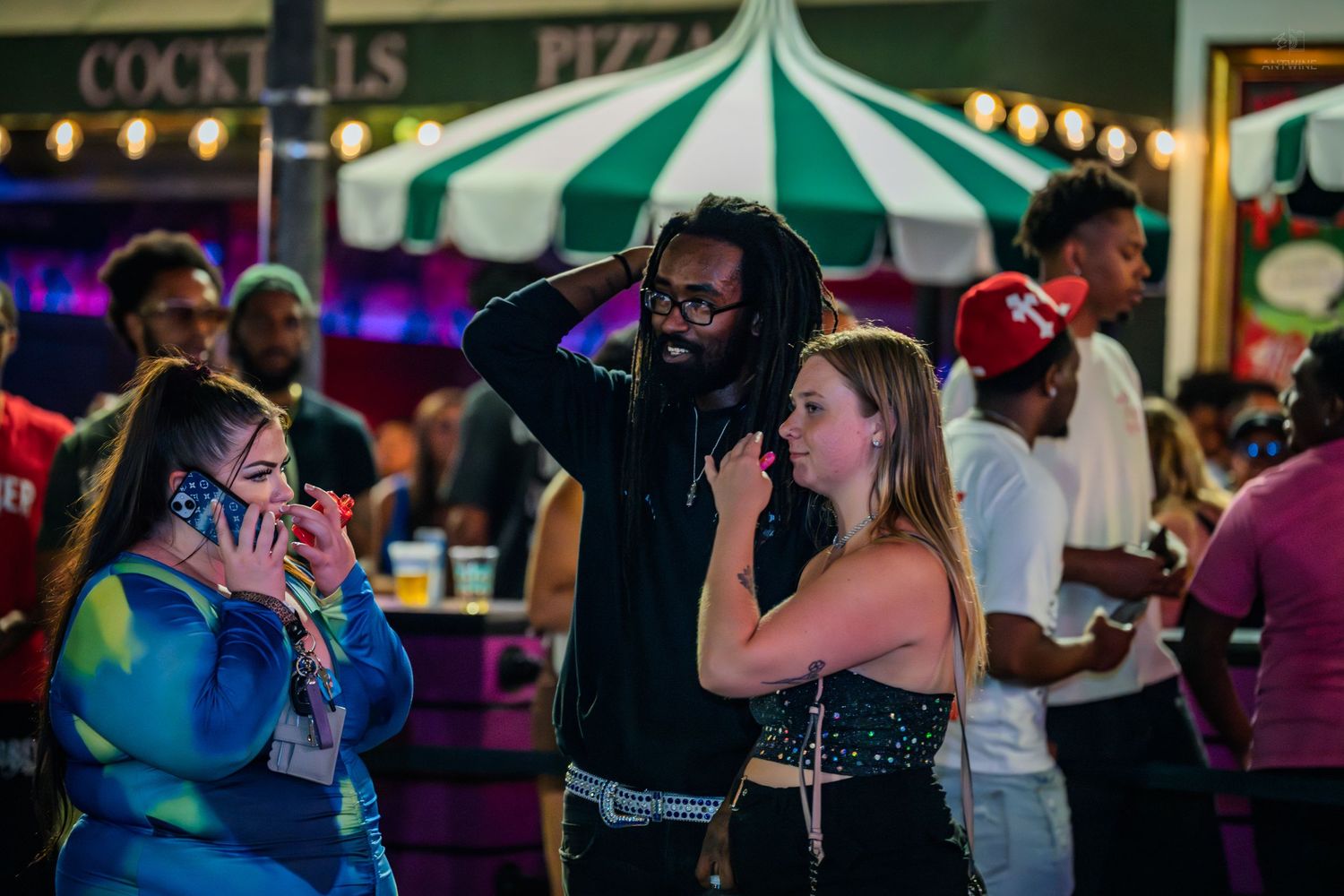 Three friends socialize at an outdoor festival near a cocktails and pizza stand.