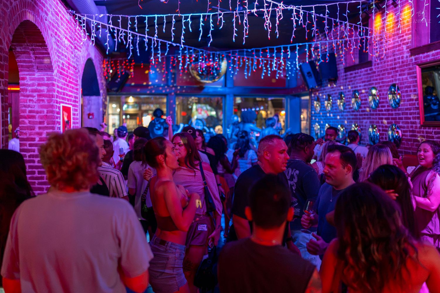 Crowded nightclub with brick walls, colorful neon lights, and people socializing on a lively dance floor.