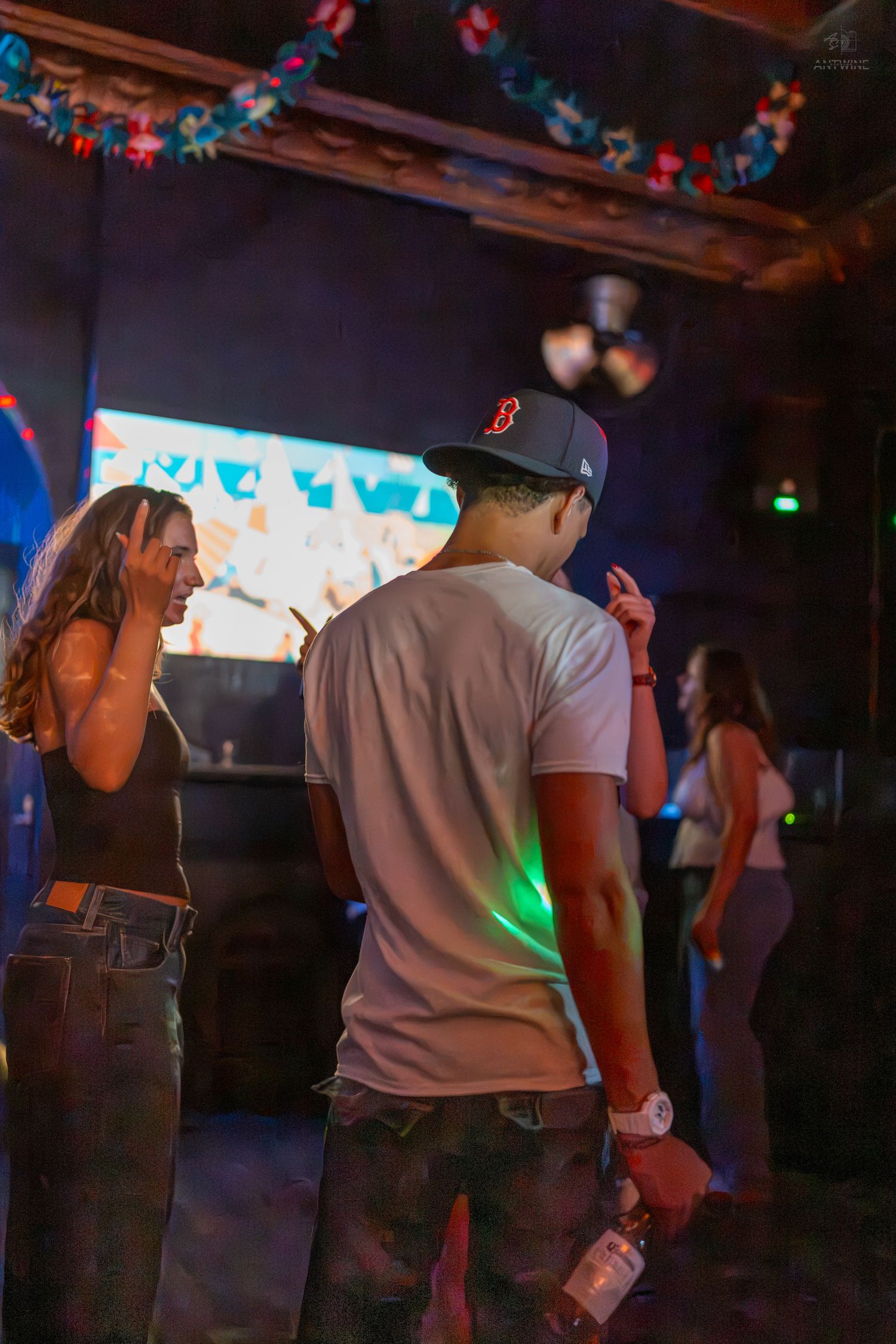 People dancing in a dimly lit club with colorful lights, a man in a Red Sox cap holds a drink.