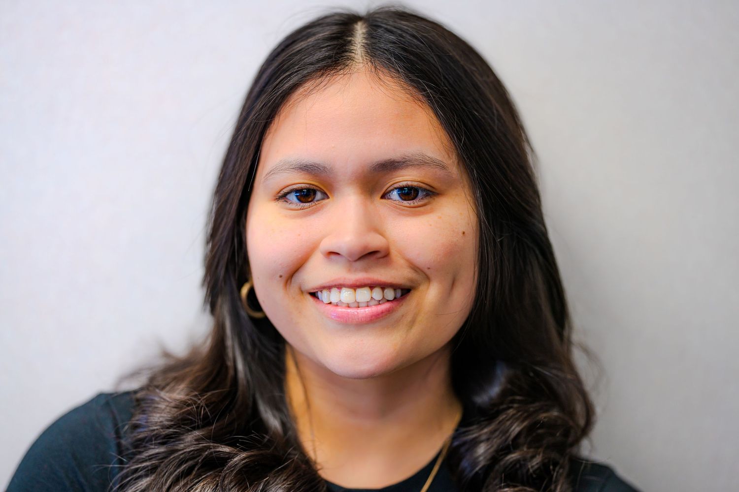 Young woman with long dark hair smiling warmly against a light gray background in a professional headshot.