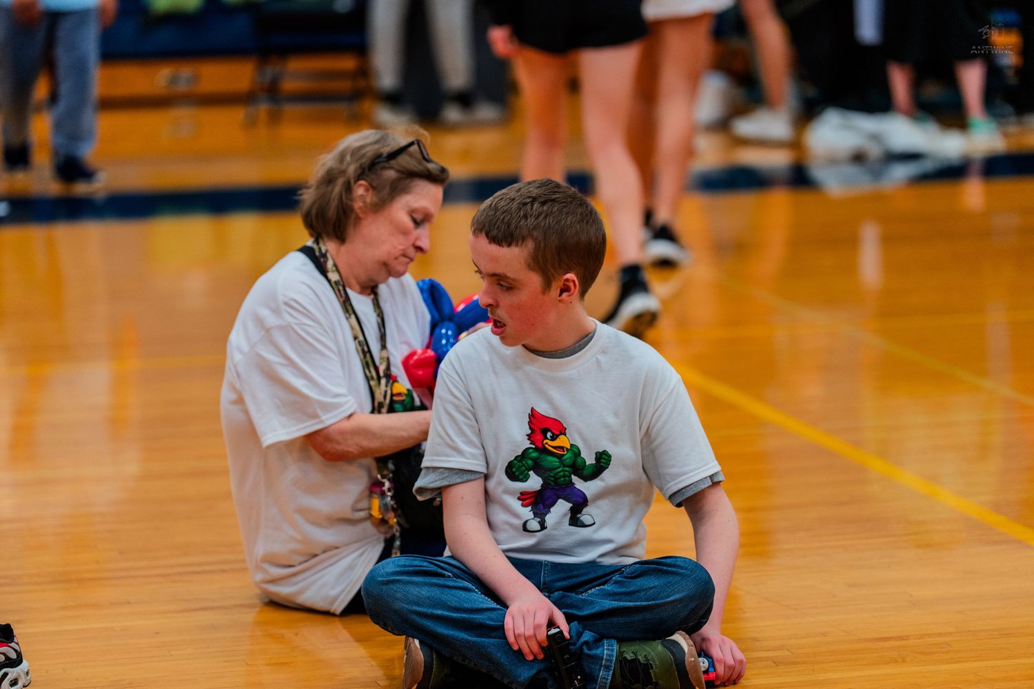 Two students sit on a gymnasium floor during a school event, one wearing a cartoon mascot graphic tee.