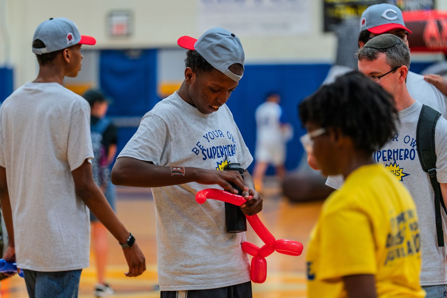 Man in superhero shirt makes balloon animal for child in gymnasium during community event.