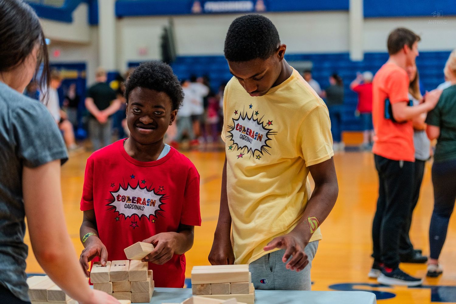 Two students smile while building with wooden blocks during a STEM competition in a school gymnasium.