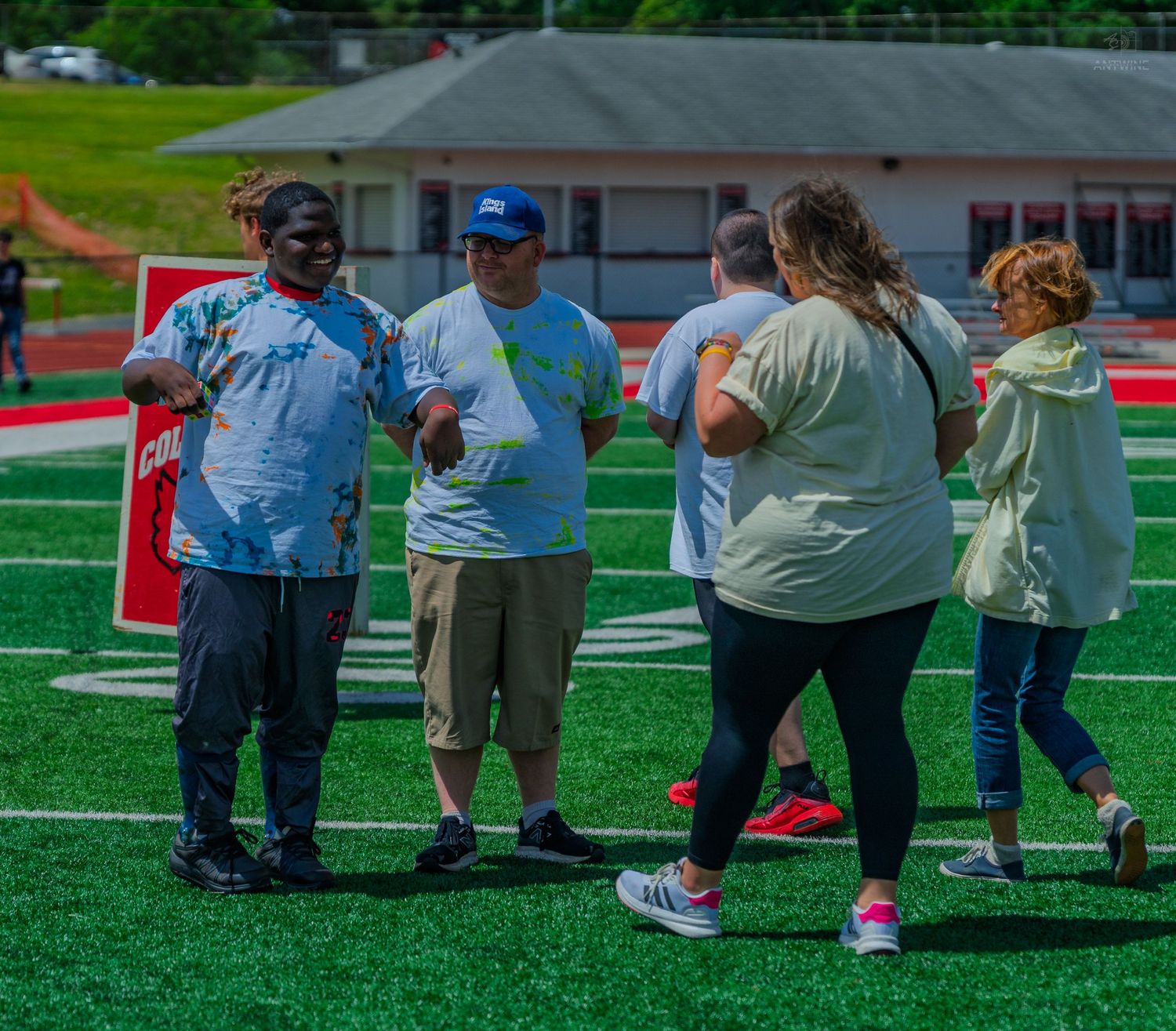 Group of adults socializing on a green turf football field during an outdoor community event on a sunny day.