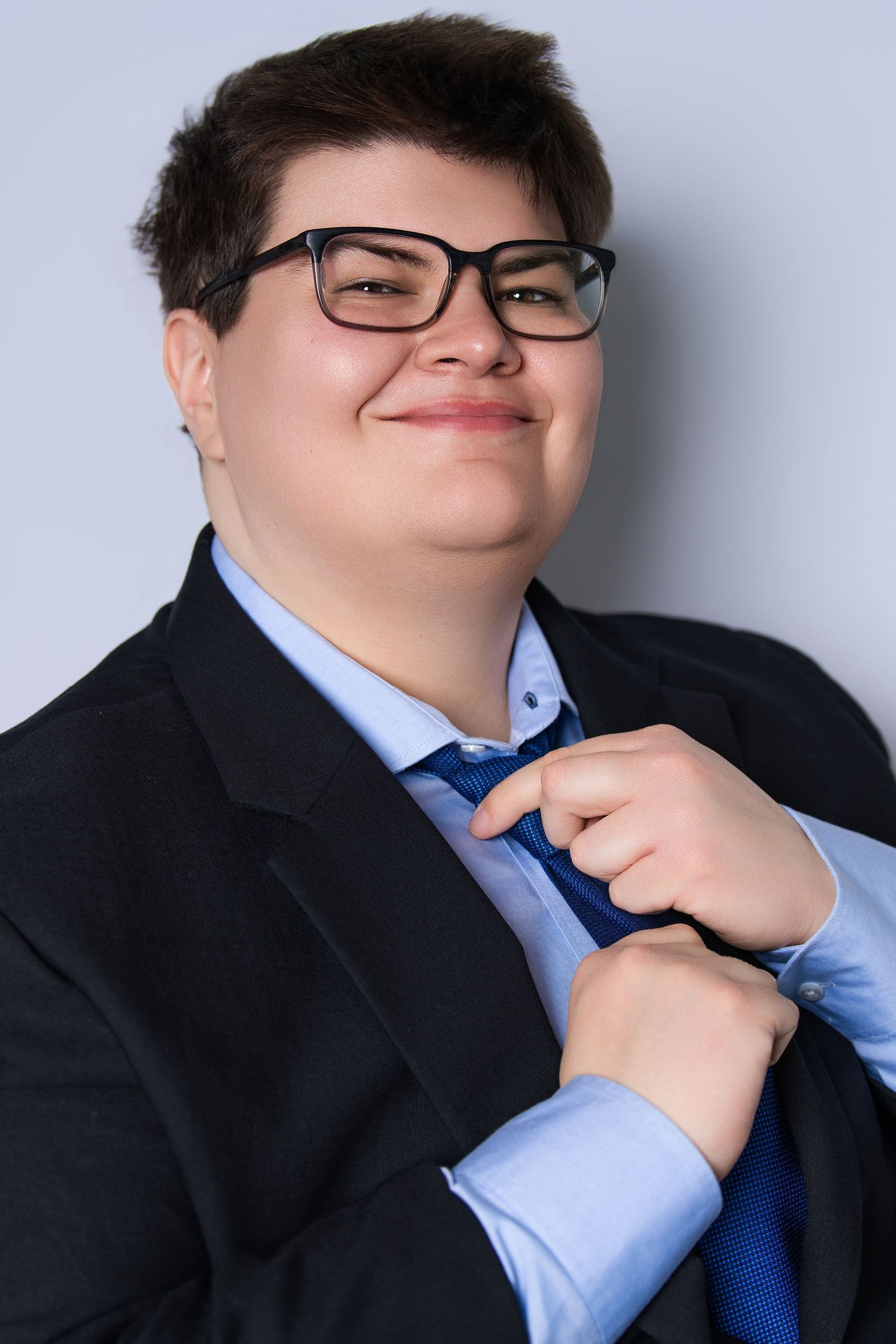 Smiling young man in black suit adjusting blue tie, wearing glasses in studio photo.