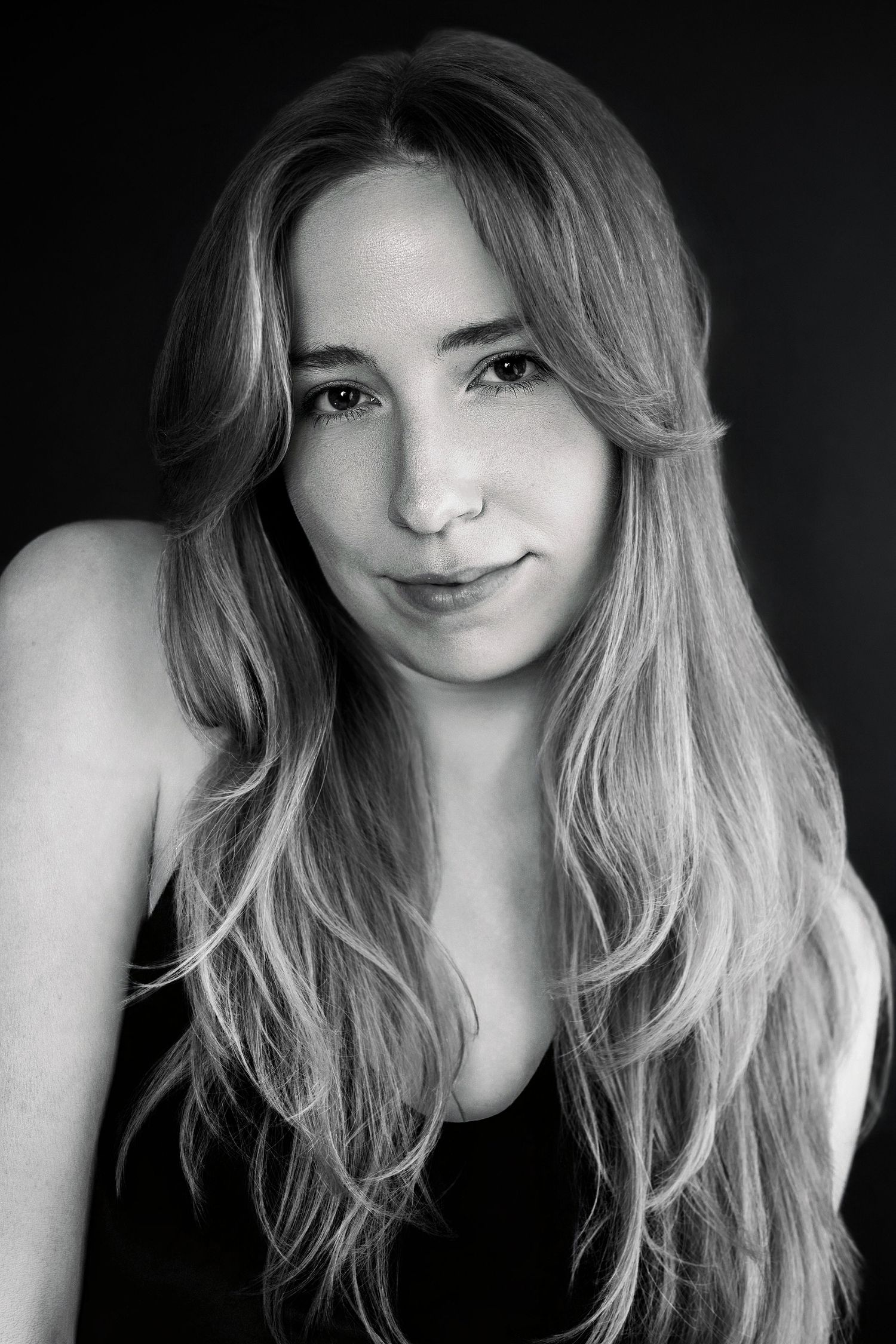 Black and white portrait of young woman with long wavy blonde hair smiling softly against dark background.