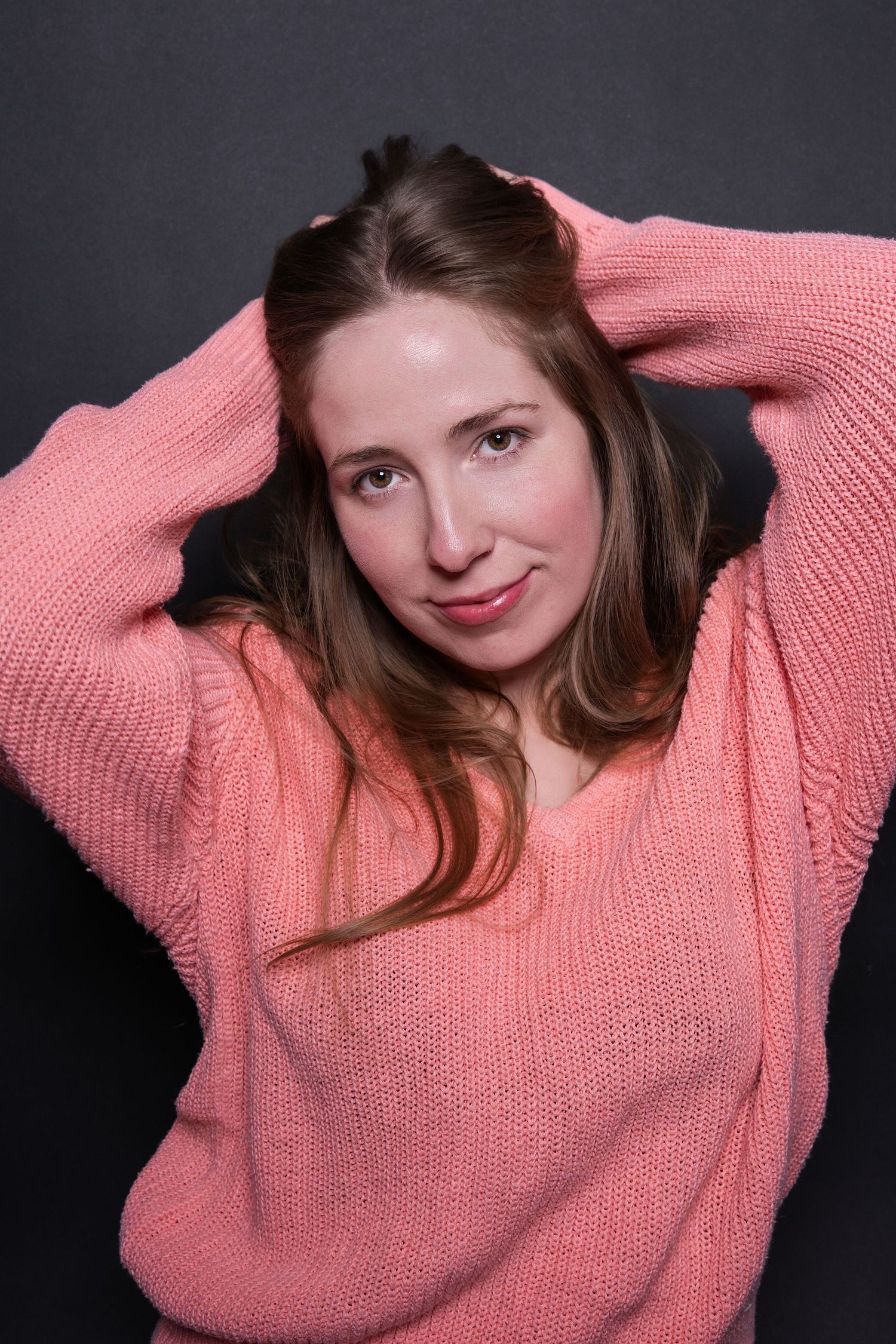 Young woman in a pink knit sweater poses with hands in hair for a studio portrait.