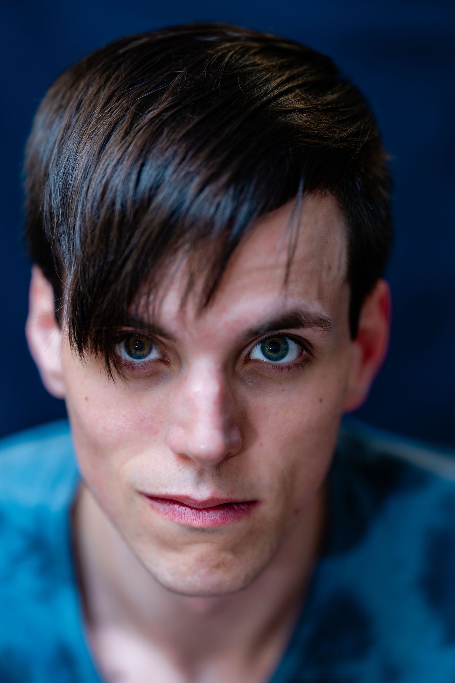 Close-up portrait of a young man with dark swept bangs and striking green eyes against a blue background.
