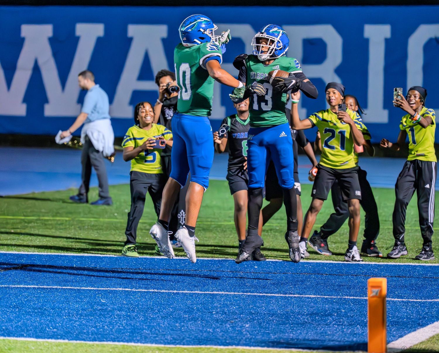High school football players celebrate a touchdown in the end zone during a night game with fans watching.