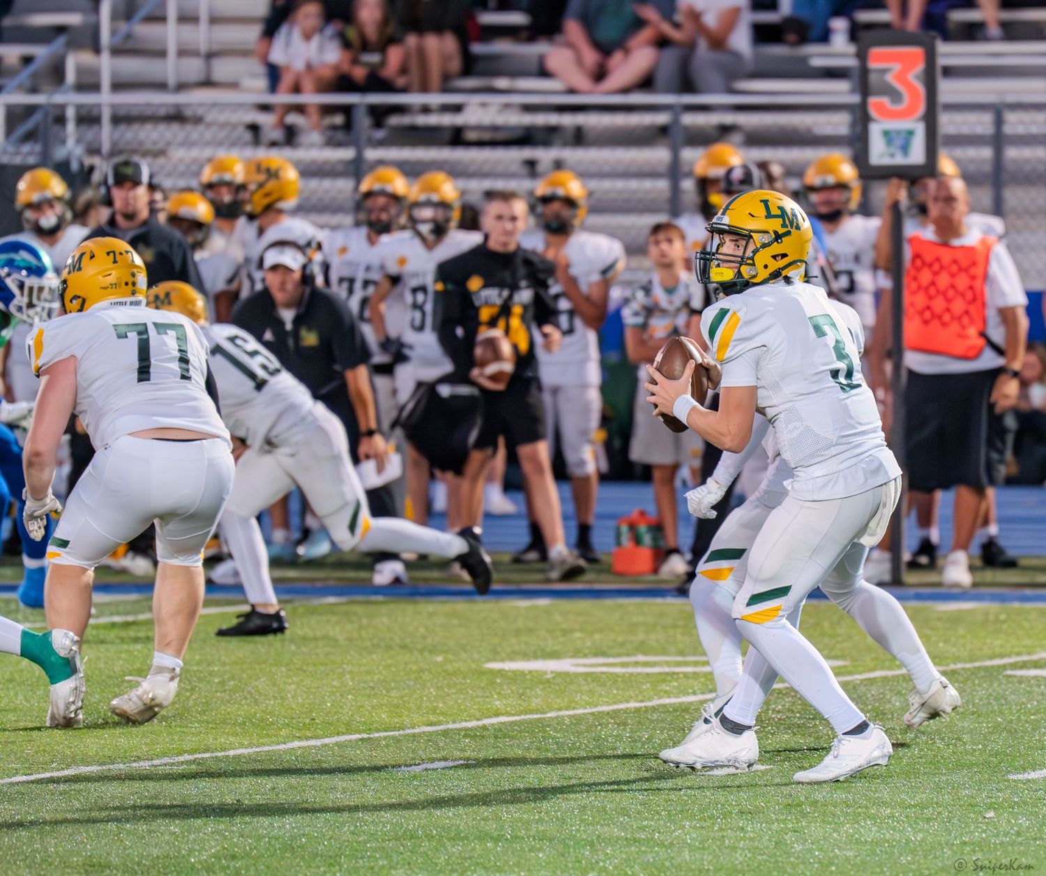 High school quarterback in white uniform drops back to pass behind offensive linemen during football game.