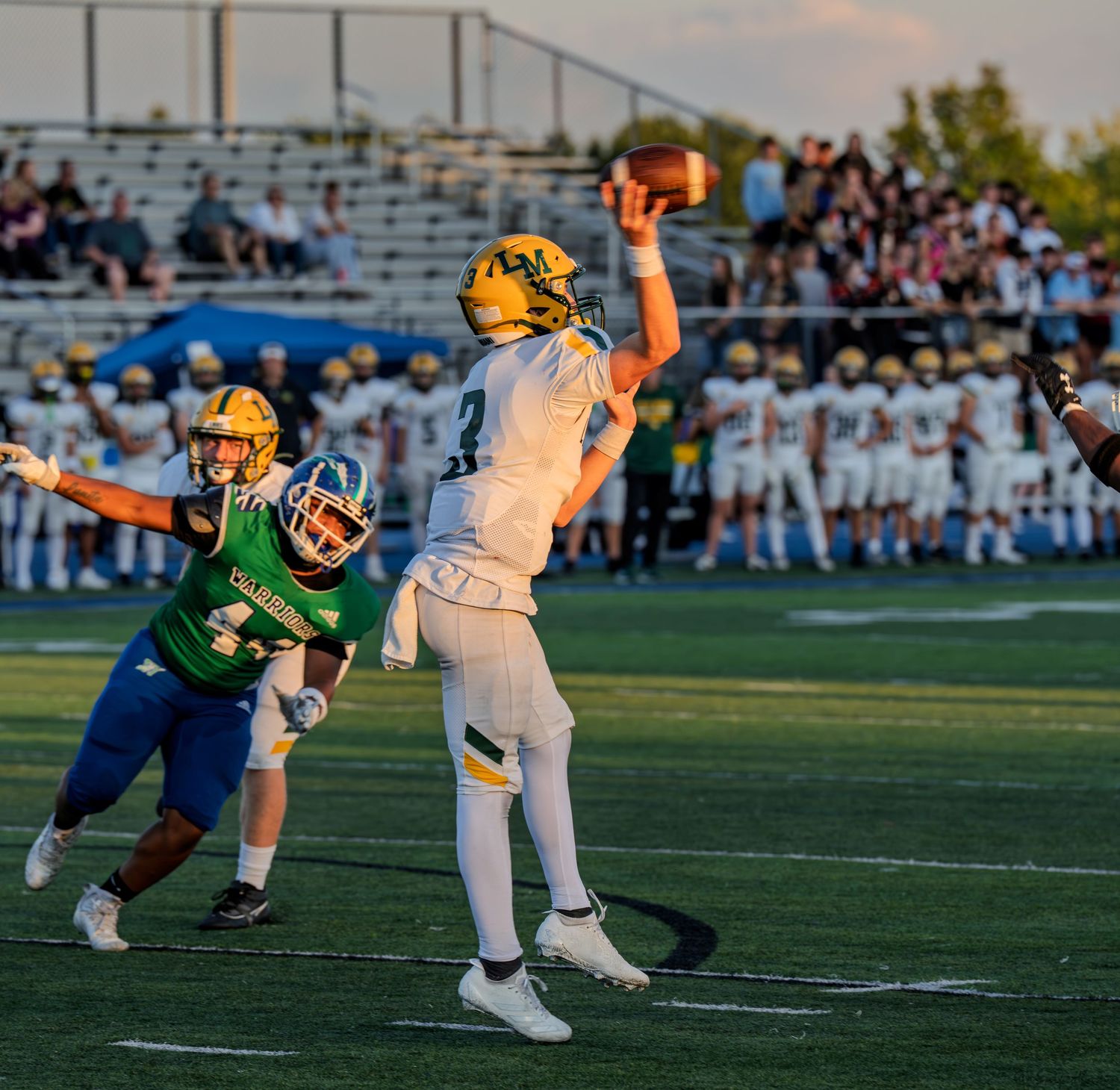 Yellow and white quarterback throws pass while Warriors defender rushes during sunny football game.