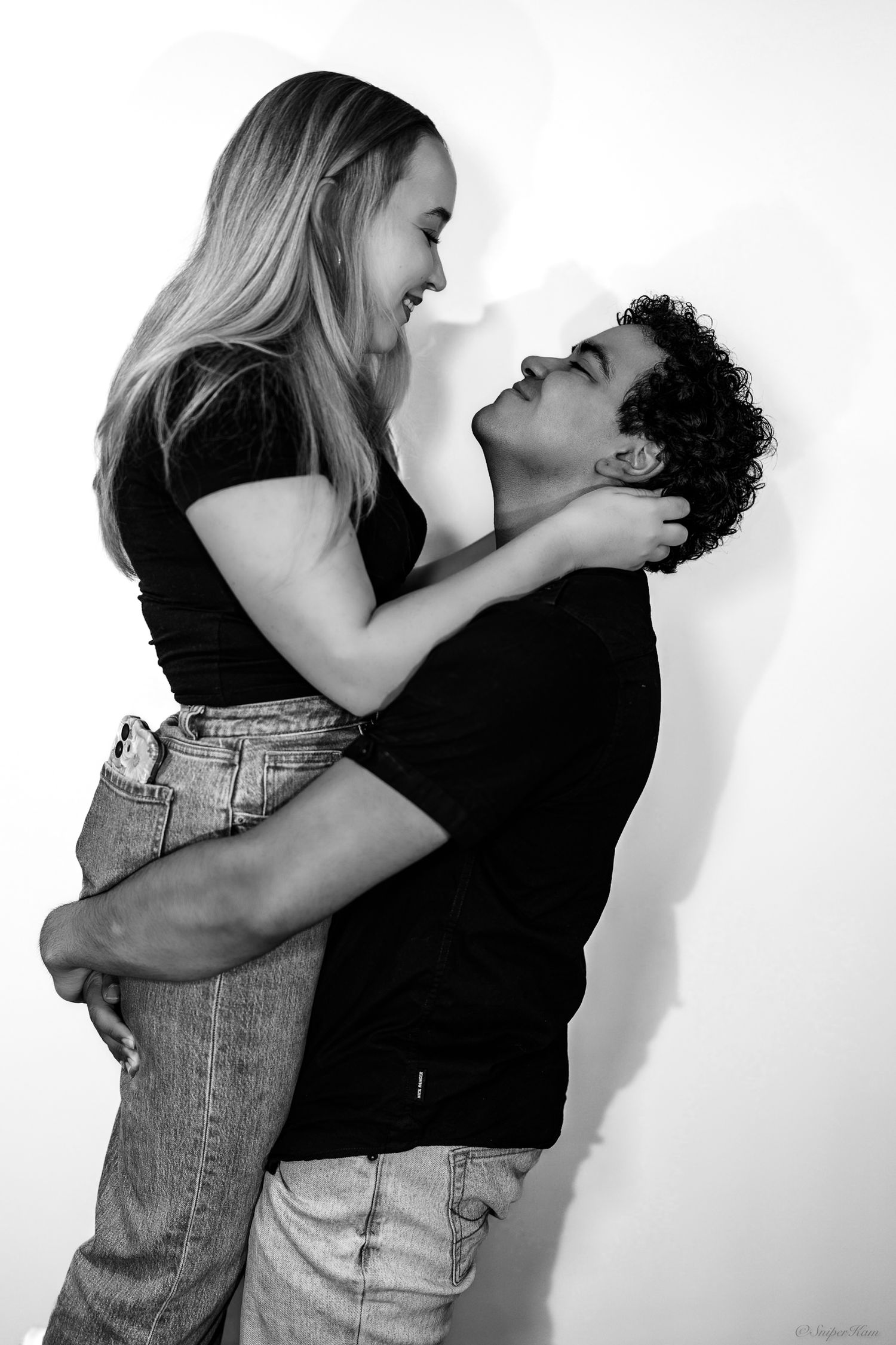 Black and white photo of couple embracing and laughing together against white wall.