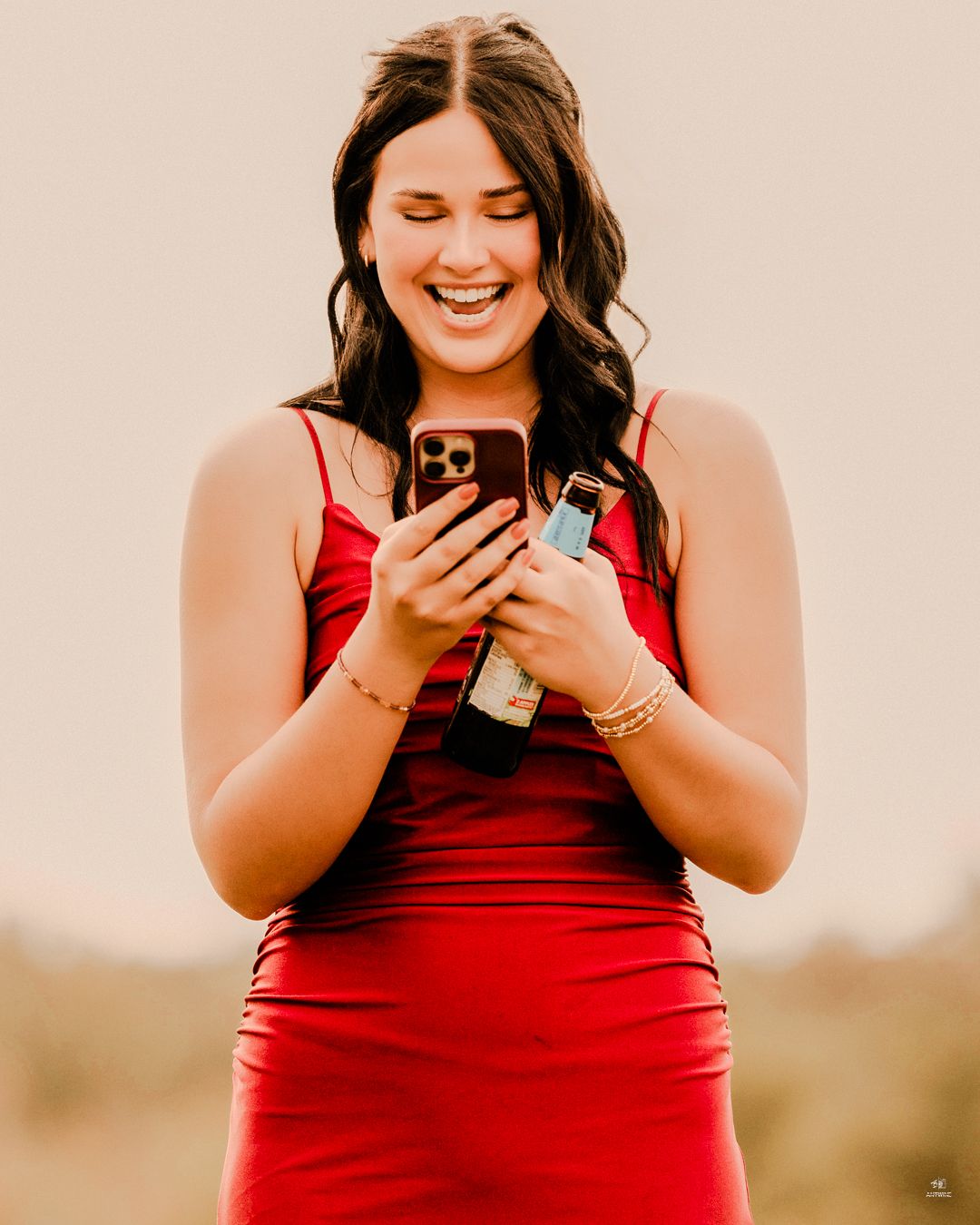 Joyful young woman in a red dress laughs while looking at her phone outdoors in warm light.