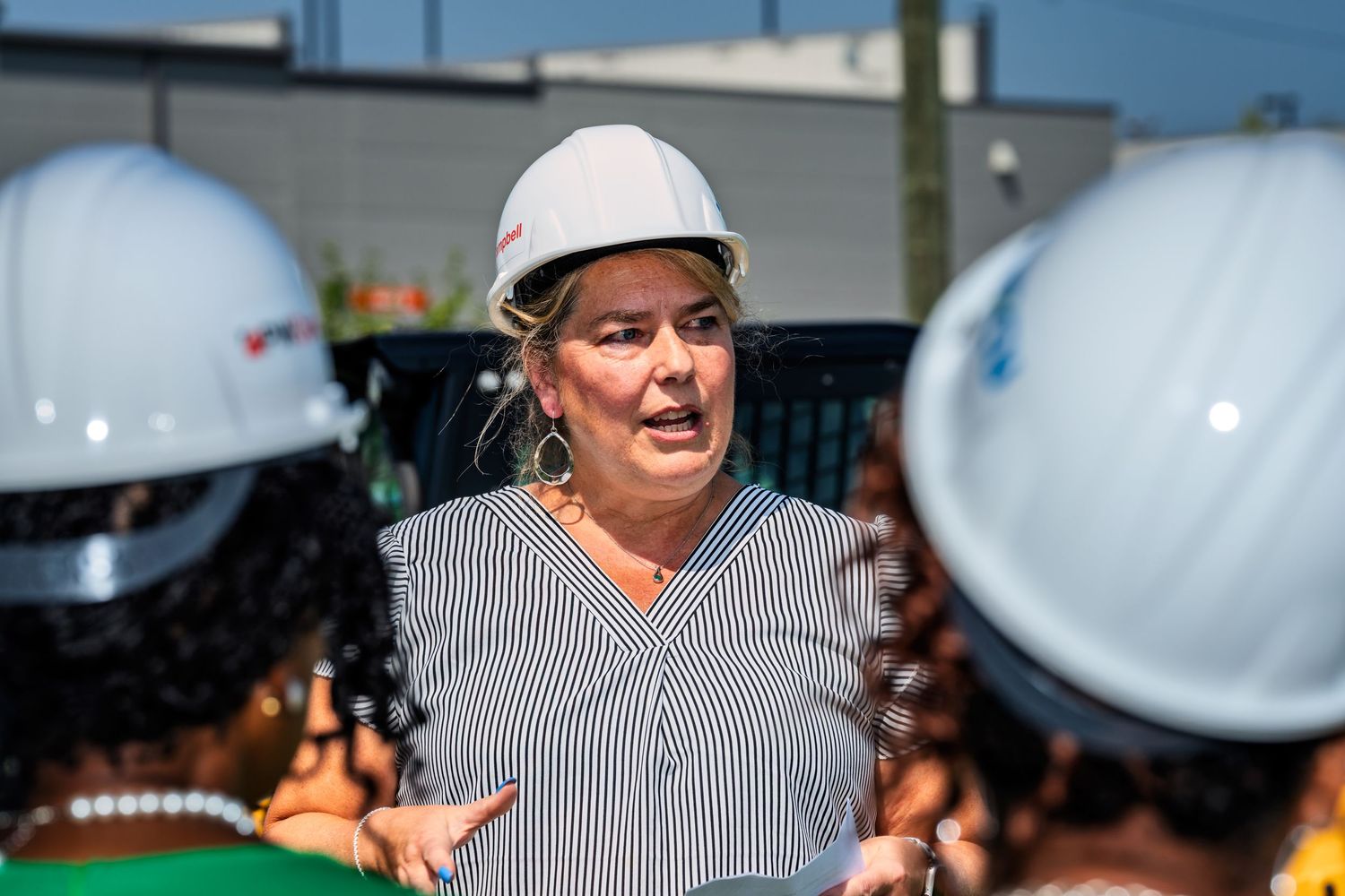 Woman in white hard hat speaking at a construction site groundbreaking event outdoors.
