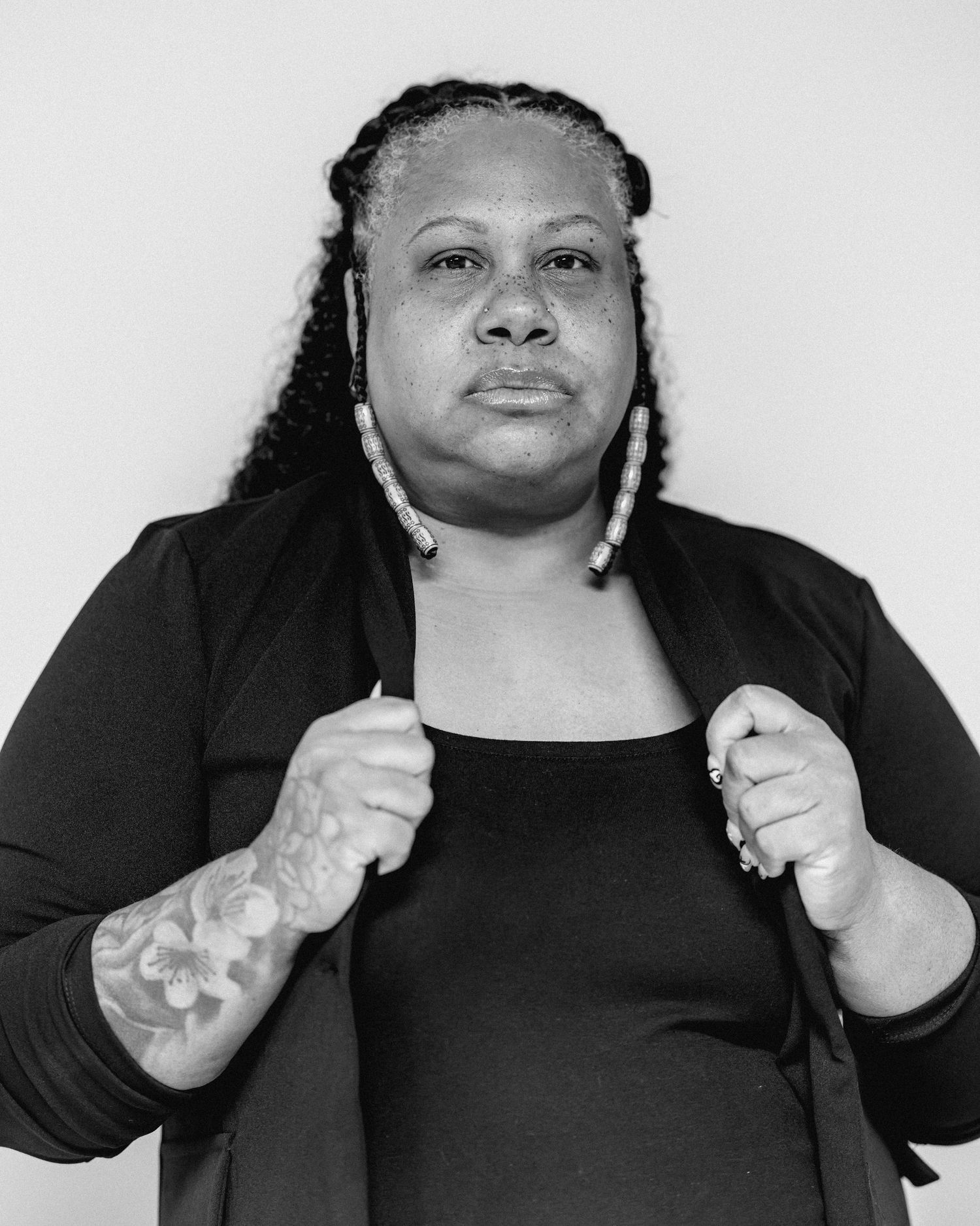 Black woman in black cardigan holding lapels with floral arm tattoo visible, monochrome portrait photo.