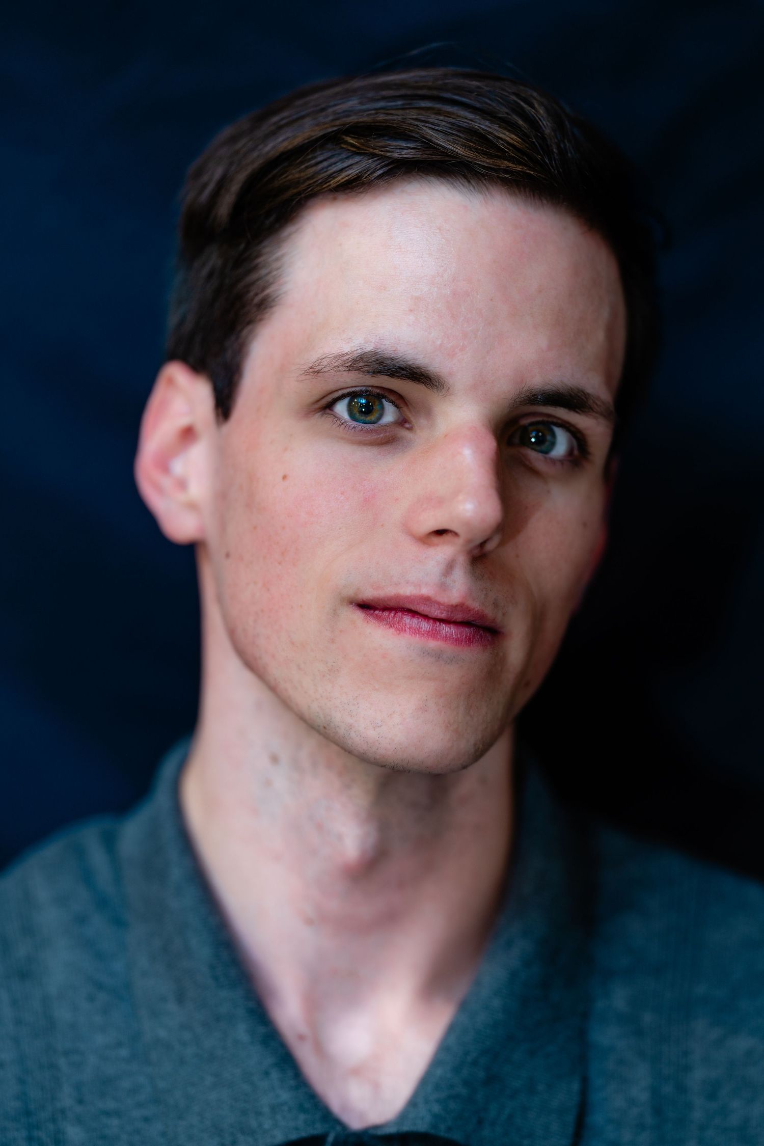 Young man with brown hair and blue-green eyes in gray polo shirt against dark blue background.
