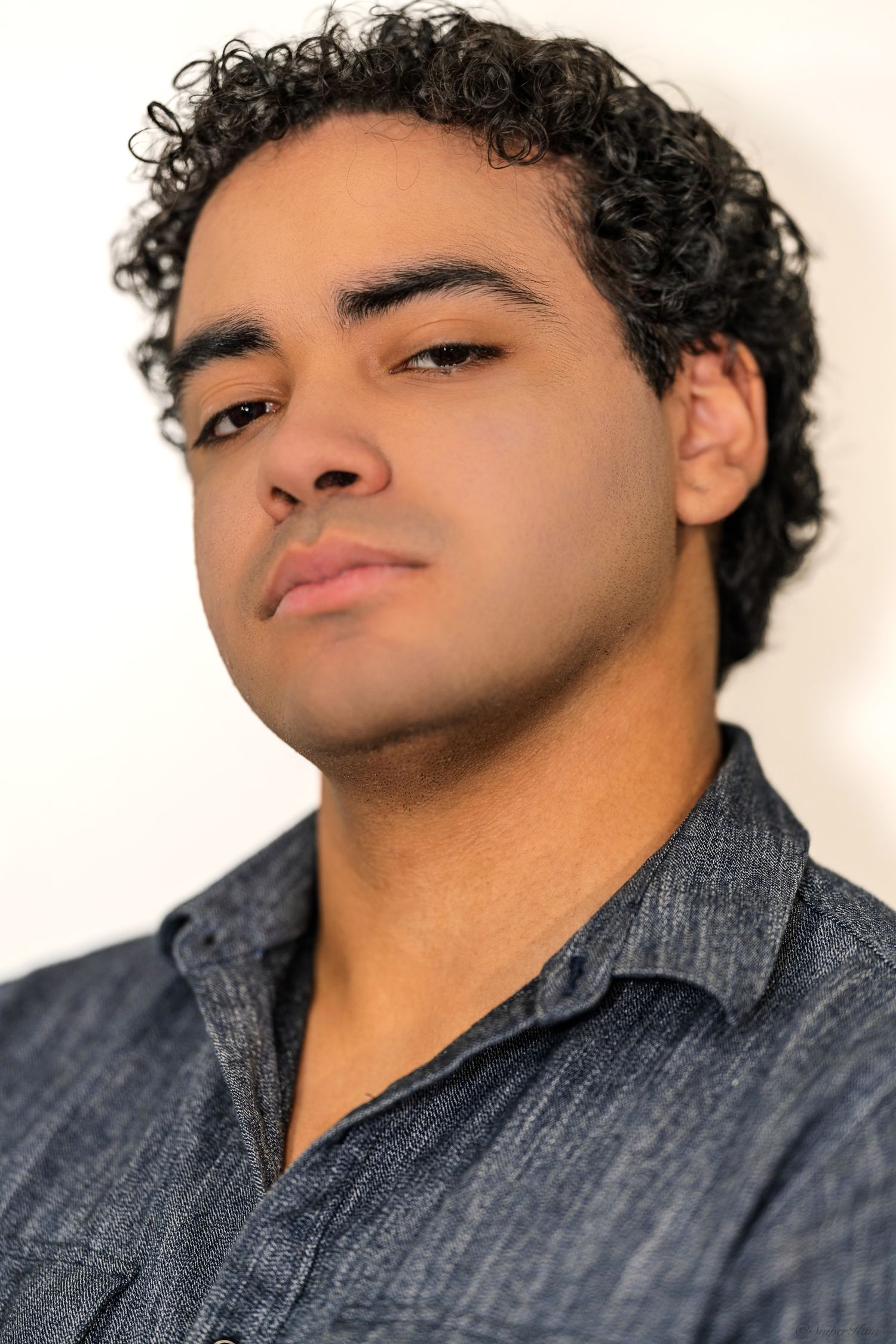 Young man with curly black hair in a blue denim shirt looking upward in a professional color headshot.