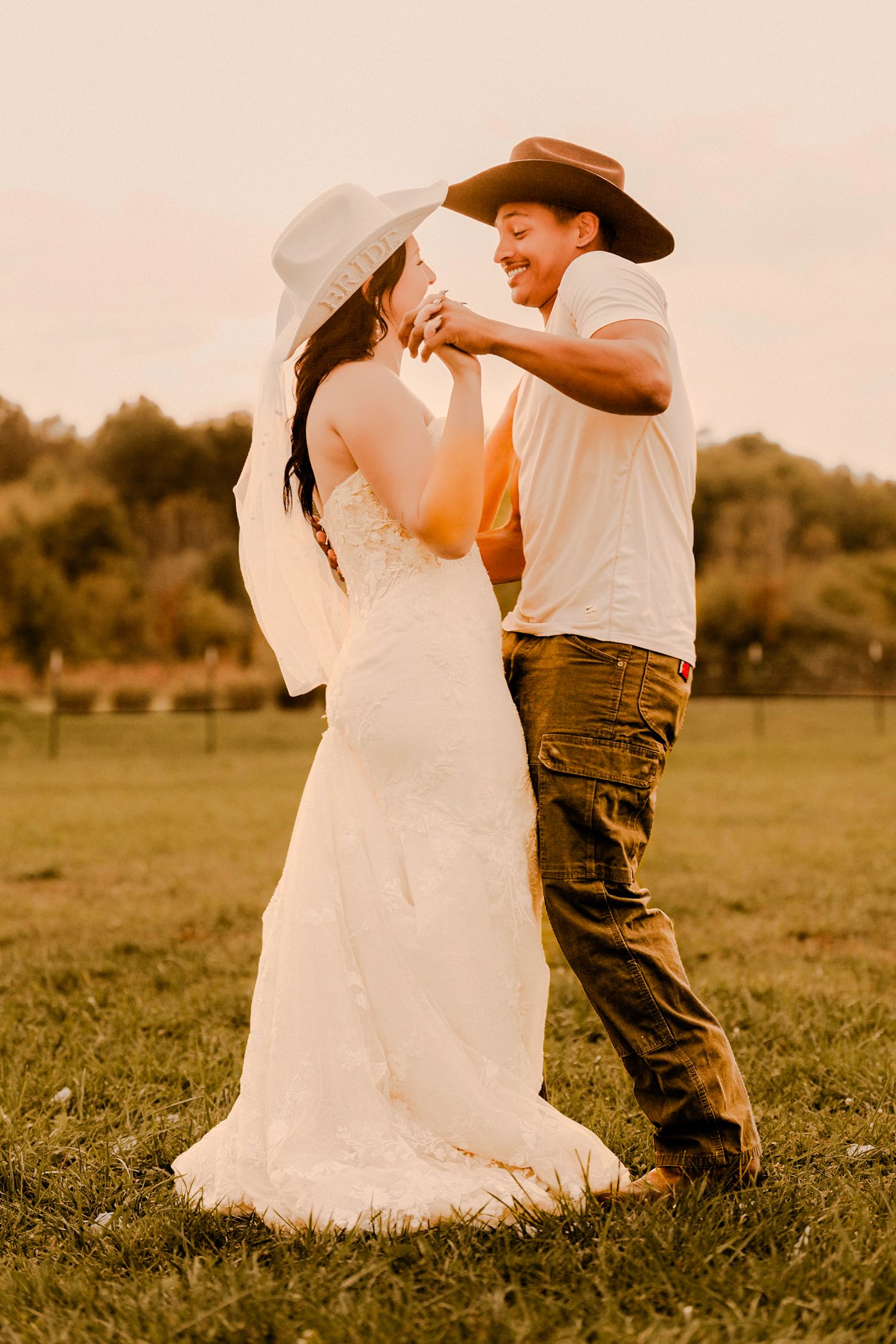 Bride and groom dancing at golden hour sunset in field, groom adjusting bride's veil.