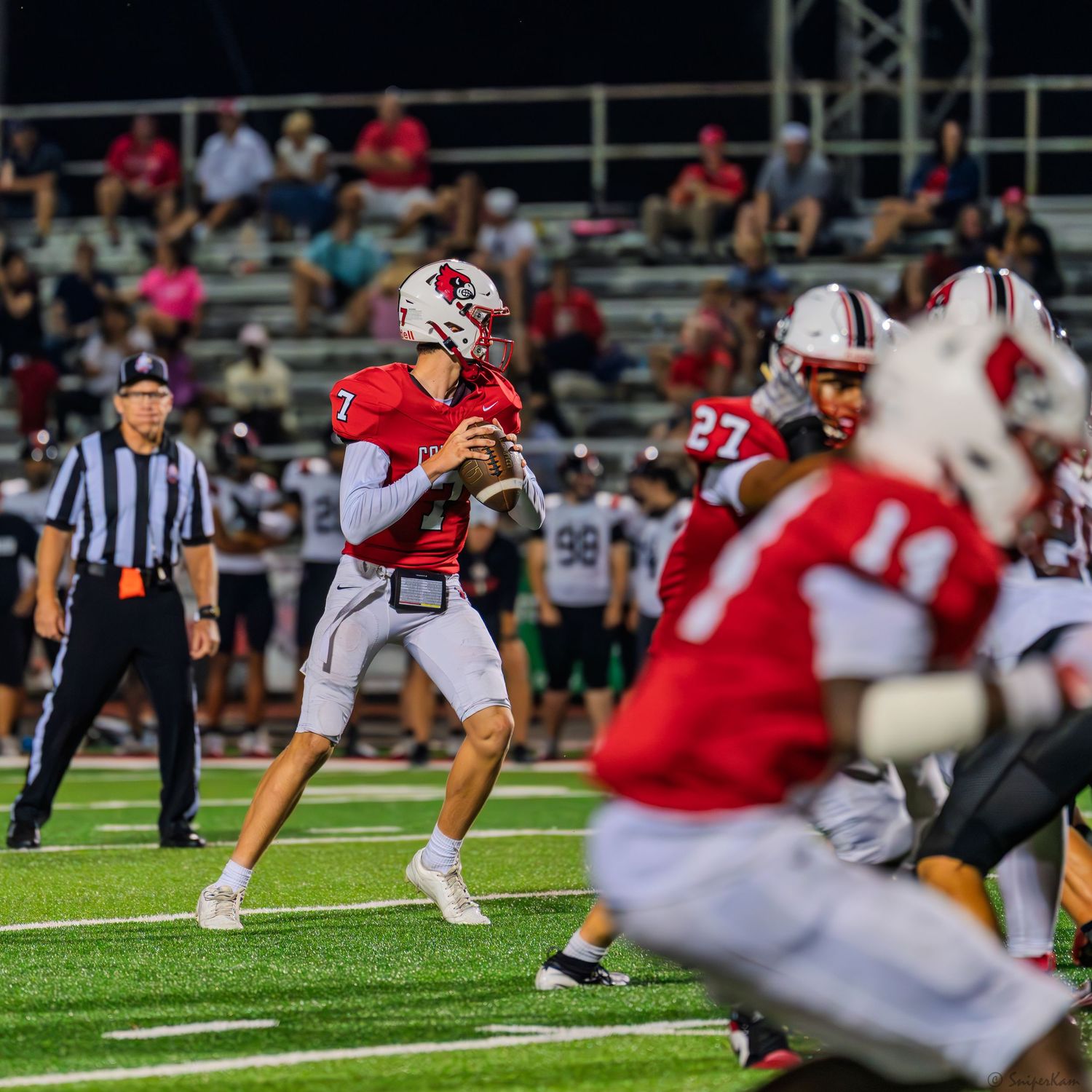 A quarterback #7 in red uniform drops back to pass with blockers protecting him during a high school football game.