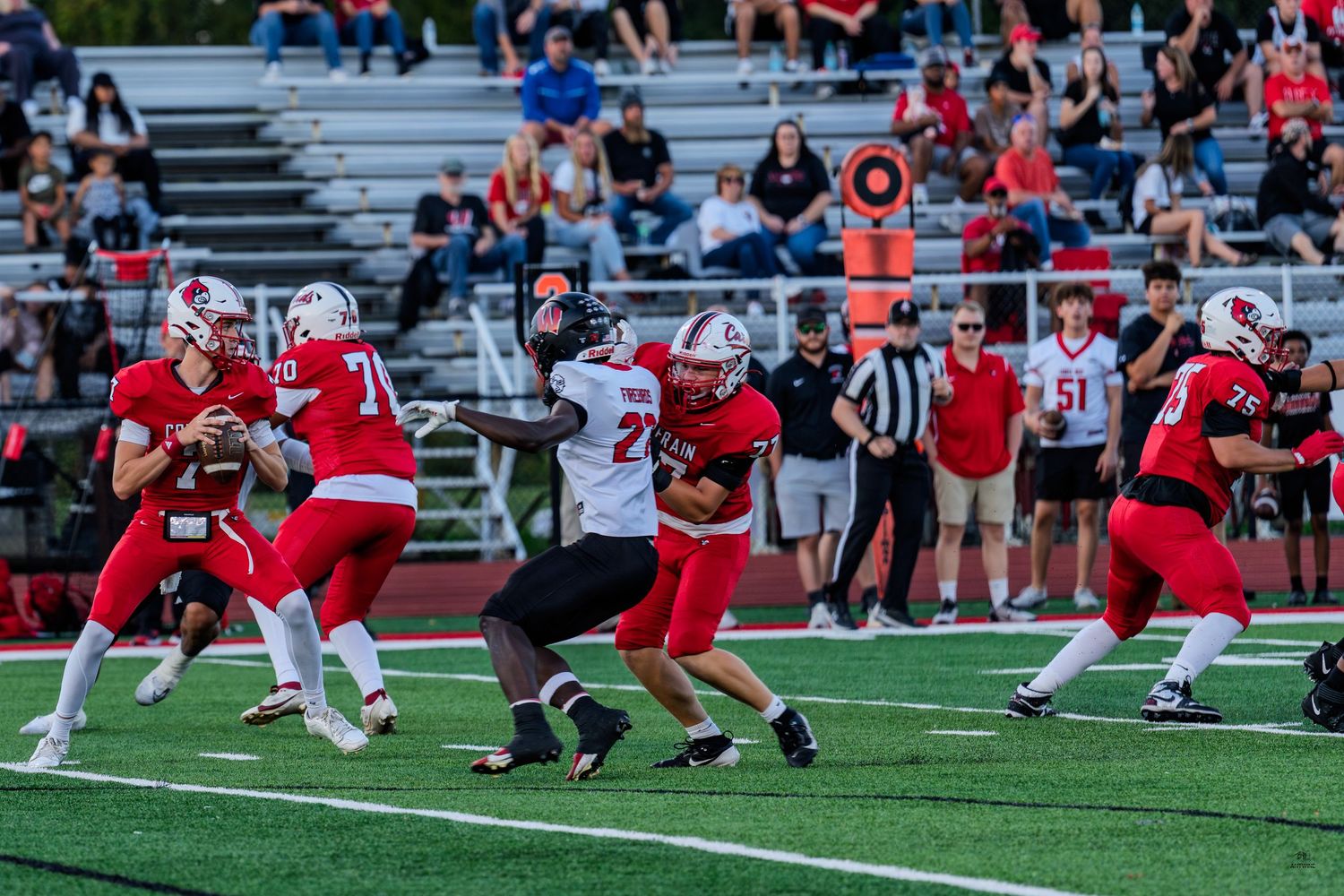 High school football quarterback in red jersey prepares to throw as defenders rush during a competitive game.