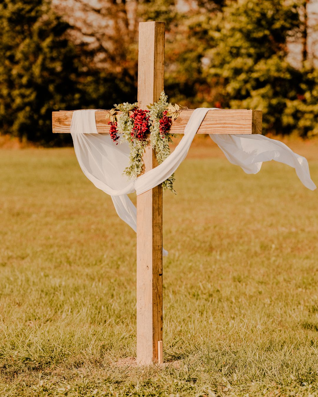 Rustic wooden cross decorated with red flowers and white fabric draping in an outdoor wedding ceremony.