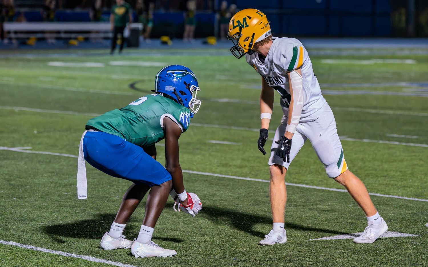 A football player in green uniform crouches in a defensive stance facing an opposing player in white and gold uniform.