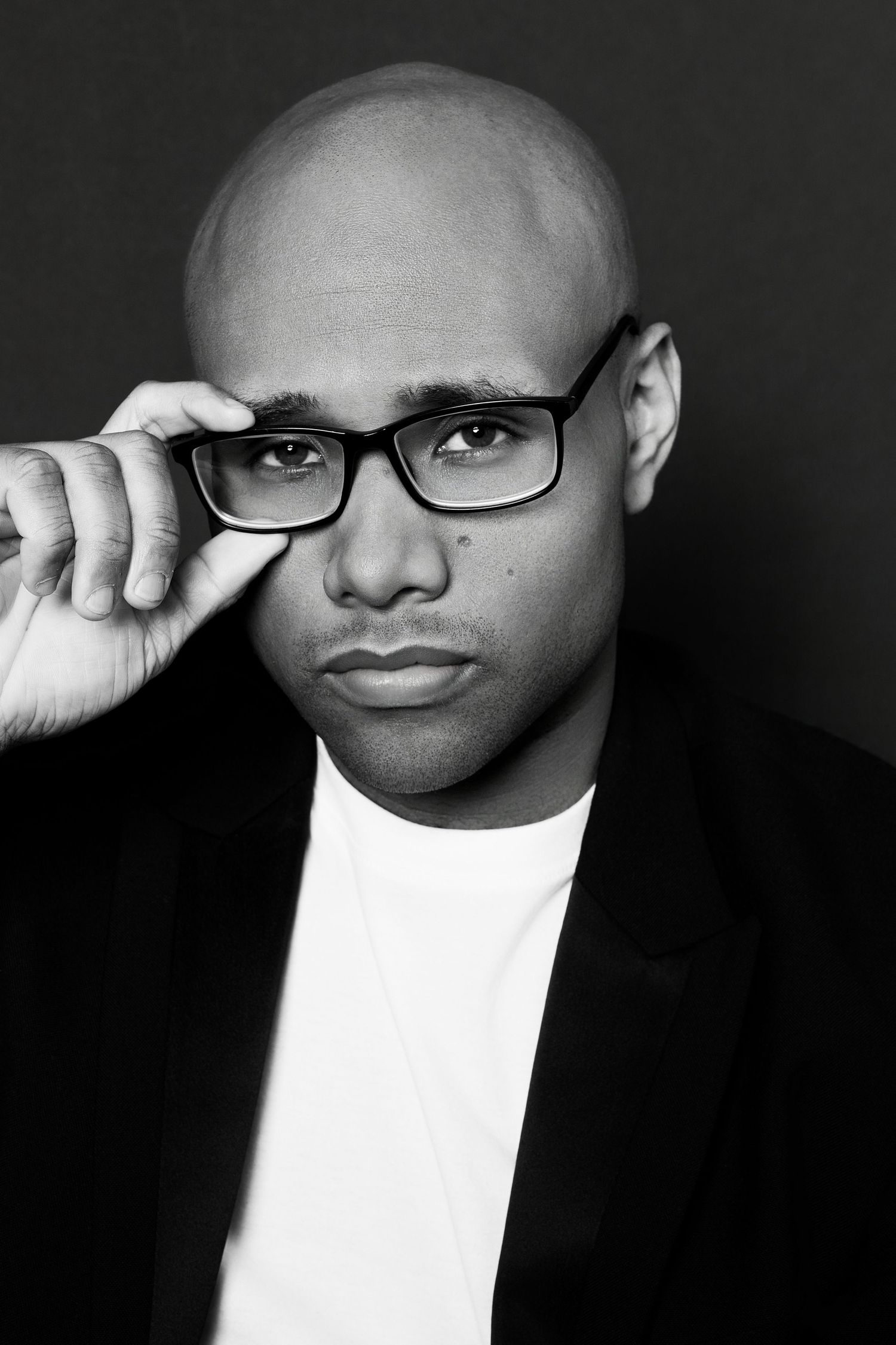 Bald Black man in black blazer adjusting rectangular glasses, serious expression in black and white portrait photo.