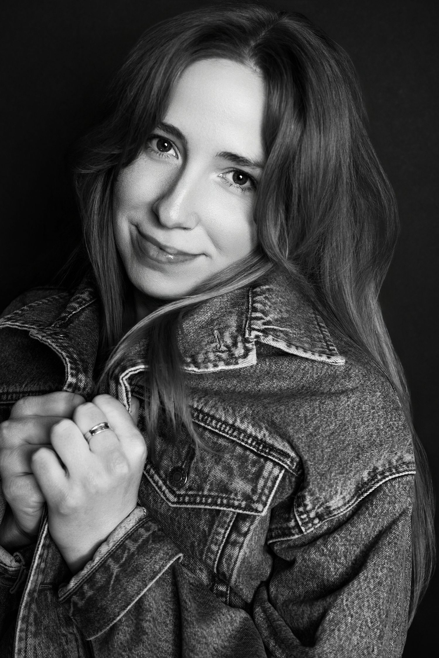 Black and white portrait of smiling young woman in denim jacket holding lapels against dark background.