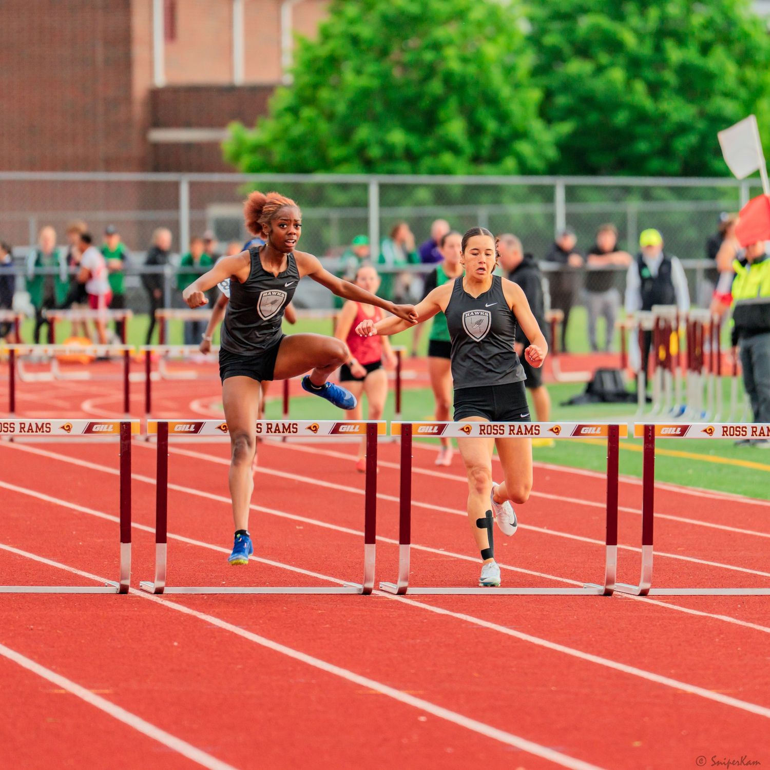 Two female track athletes in black uniforms clear hurdles together on a Ross Rams red track.