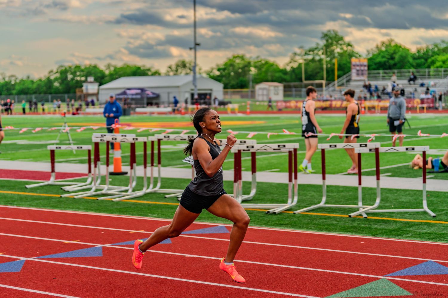 Female track athlete sprints powerfully past hurdles on a red track during a competitive meet.