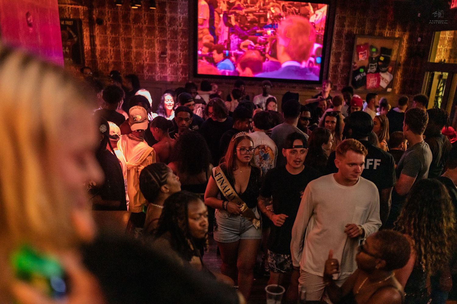 Lively bar scene with crowd of young adults partying under pink neon lights and large video screen.