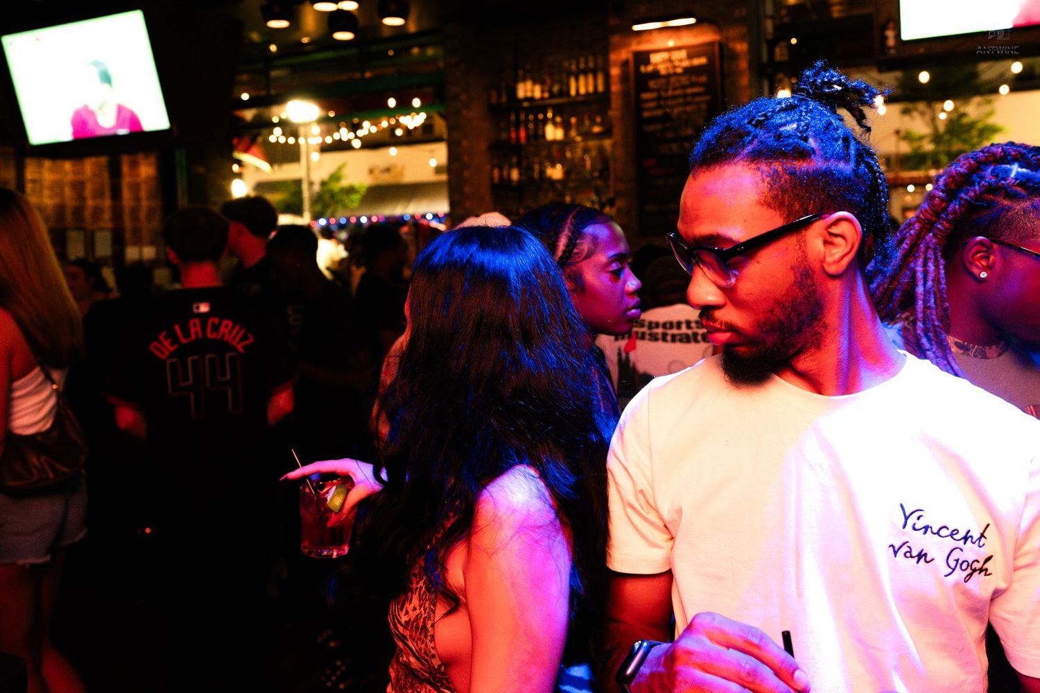 Man in Vincent Van Gogh shirt socializing at a crowded nightclub bar event.