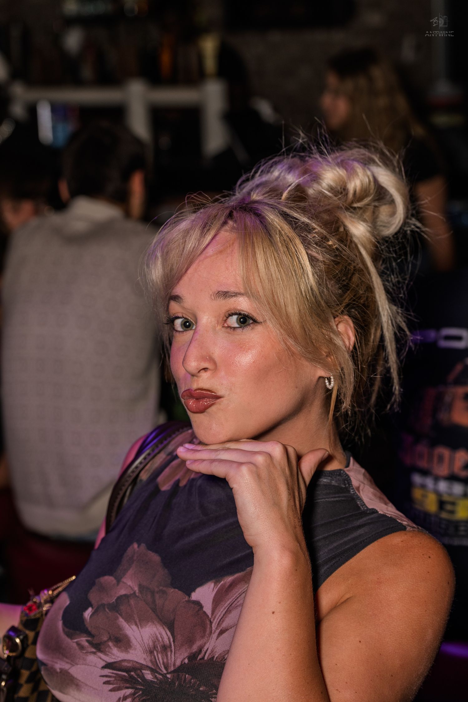 Young blonde woman with purple-highlighted updo posing thoughtfully at a lively nightclub venue.