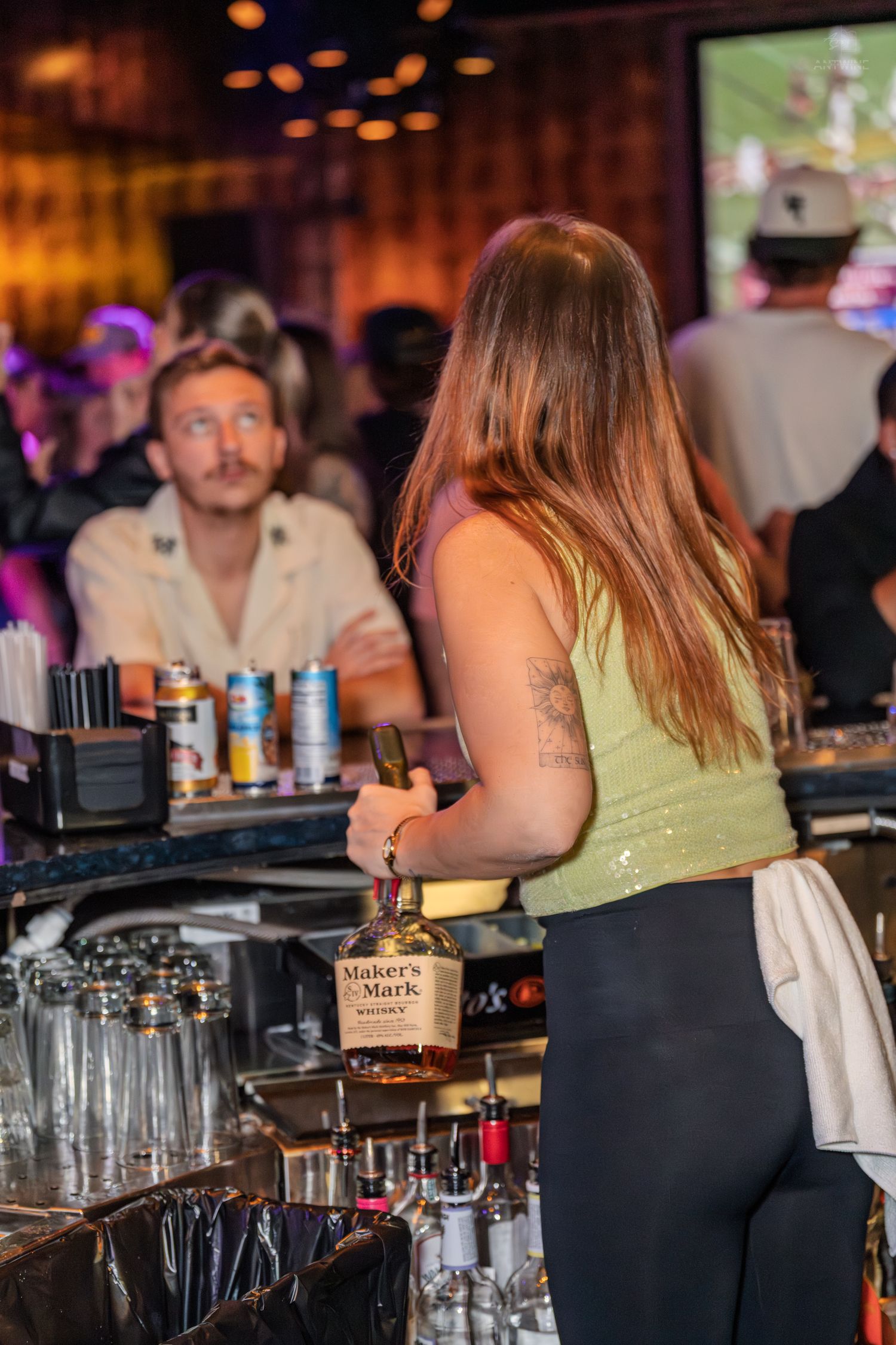 Female bartender holding a Maker's Mark whiskey bottle behind a busy bar counter.