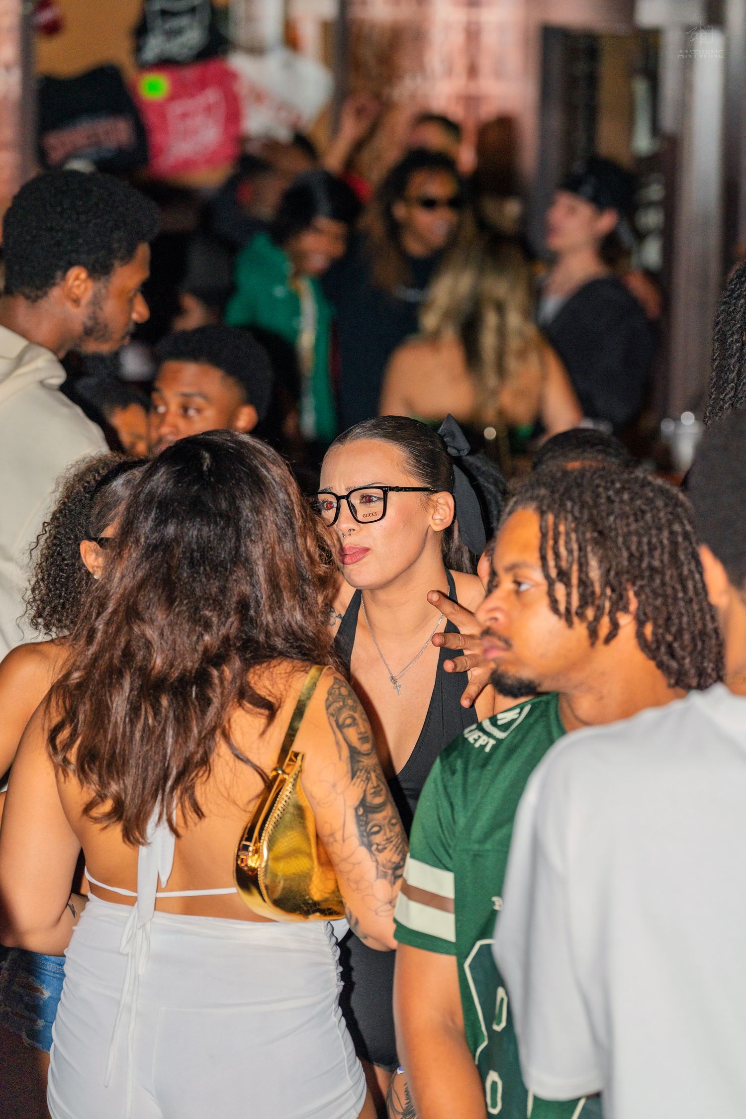 Woman with glasses and black hair bow socializing in a crowded bar or nightclub setting.