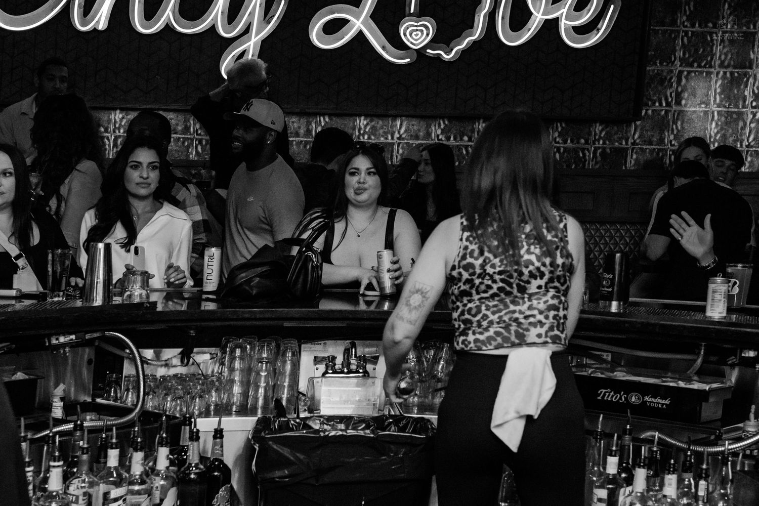 Women socializing at a bar under an Only Love neon sign with bottles on the counter.