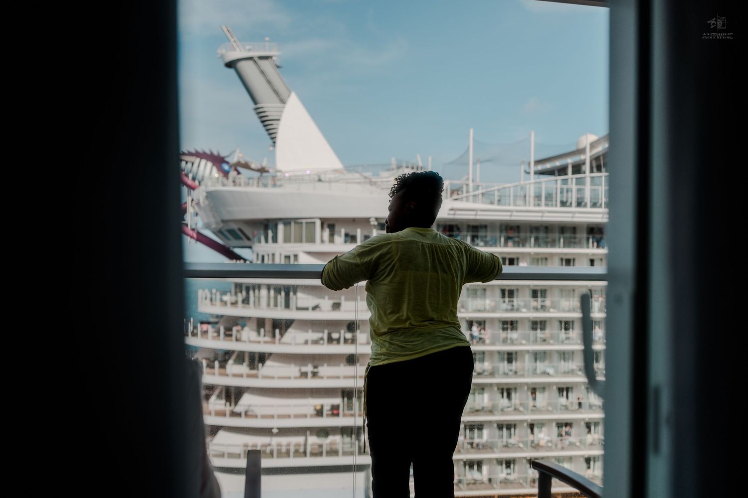 Person in green shirt gazing at a large cruise ship from a window or balcony viewpoint.