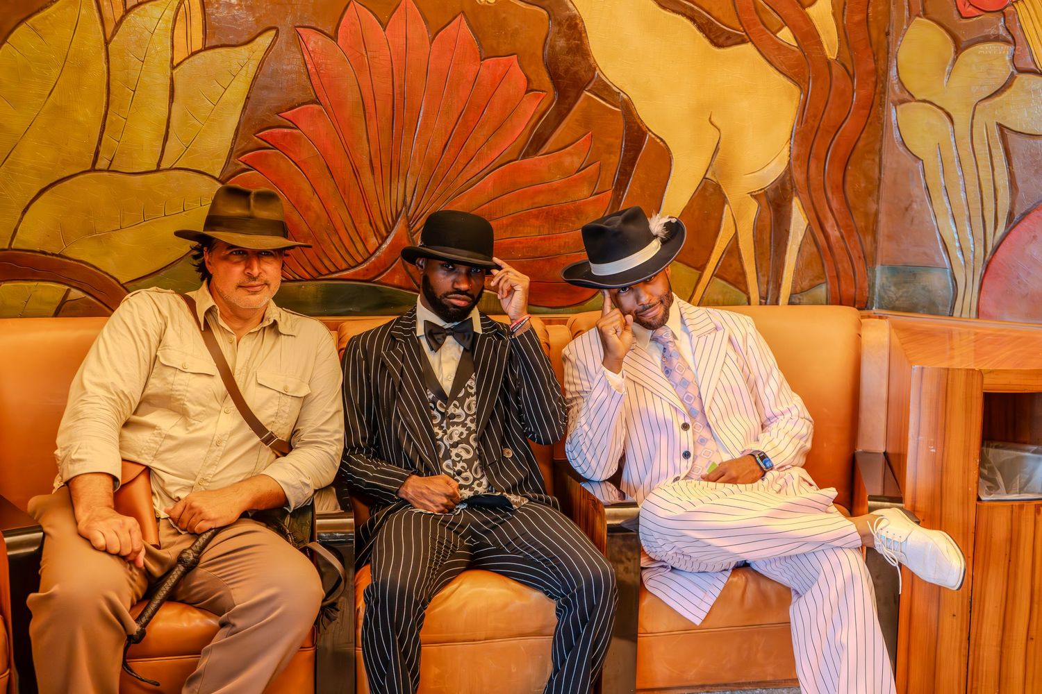 Three stylish men in vintage suits and hats pose on a couch before an Art Deco mural backdrop.