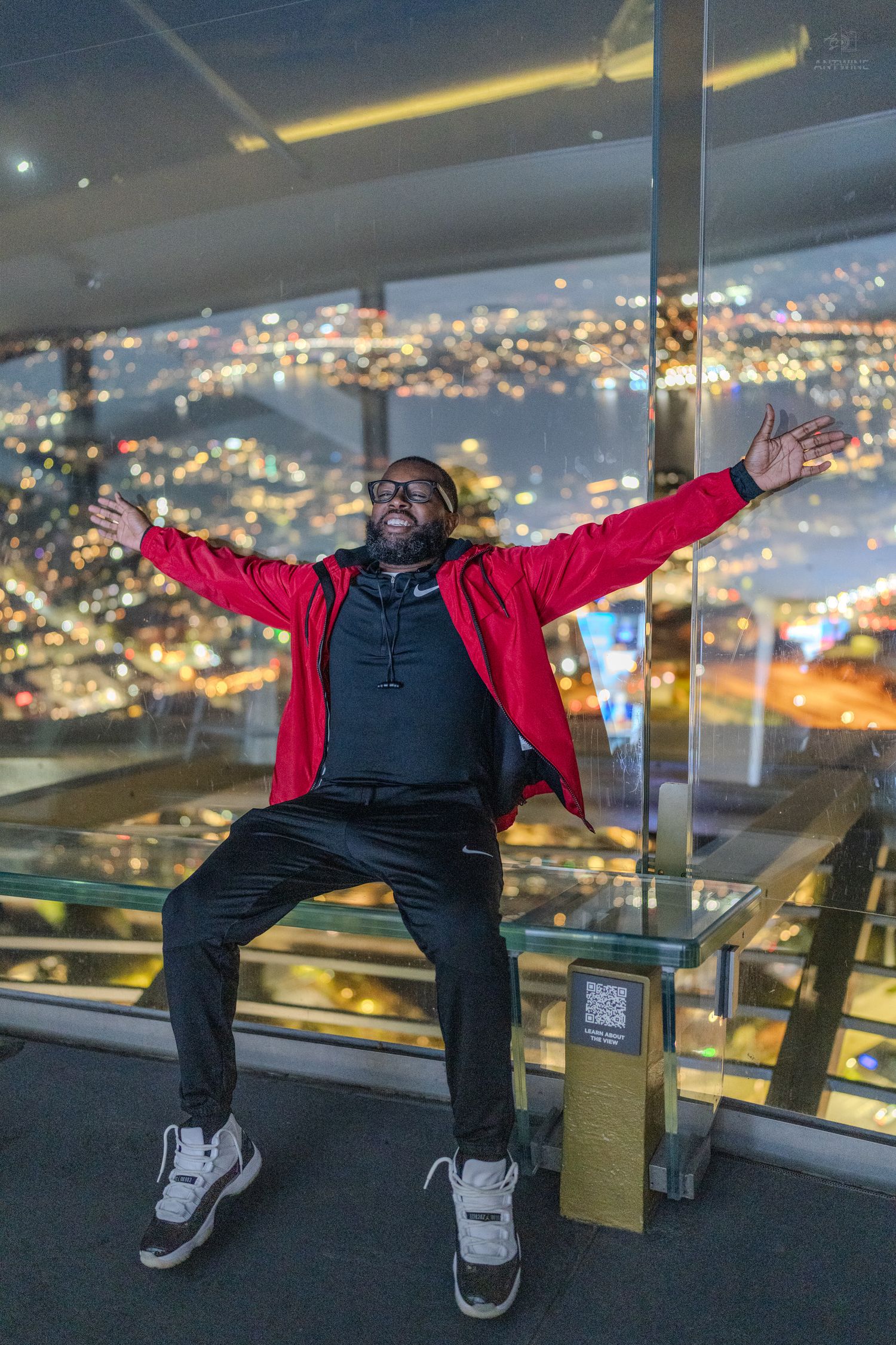A joyful man in a red jacket sits with arms outstretched on an observation deck with city lights behind him.